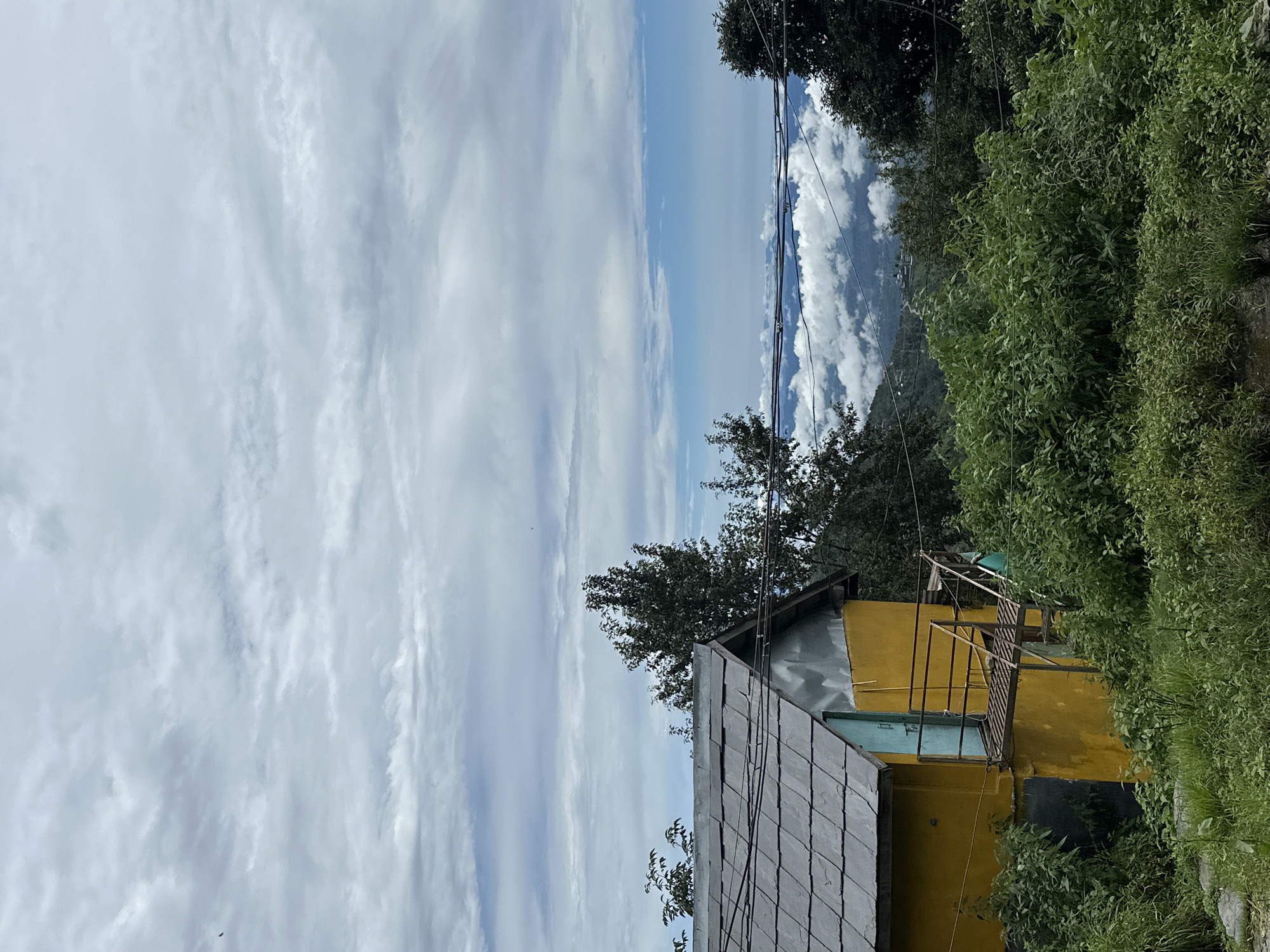 The image shows a yellow building with a grey slate roof, located in Dharamkot, India. The building has a metal walkway and stairs leading to a teal door. It is surrounded by lush green vegetation and trees. In the background, there are mountains covered in clouds, with a hint of a village nestled in the valley. The sky is cloudy, with patches of blue visible. The scene appears to be a rural or mountainous area, possibly a guesthouse or a residential building.