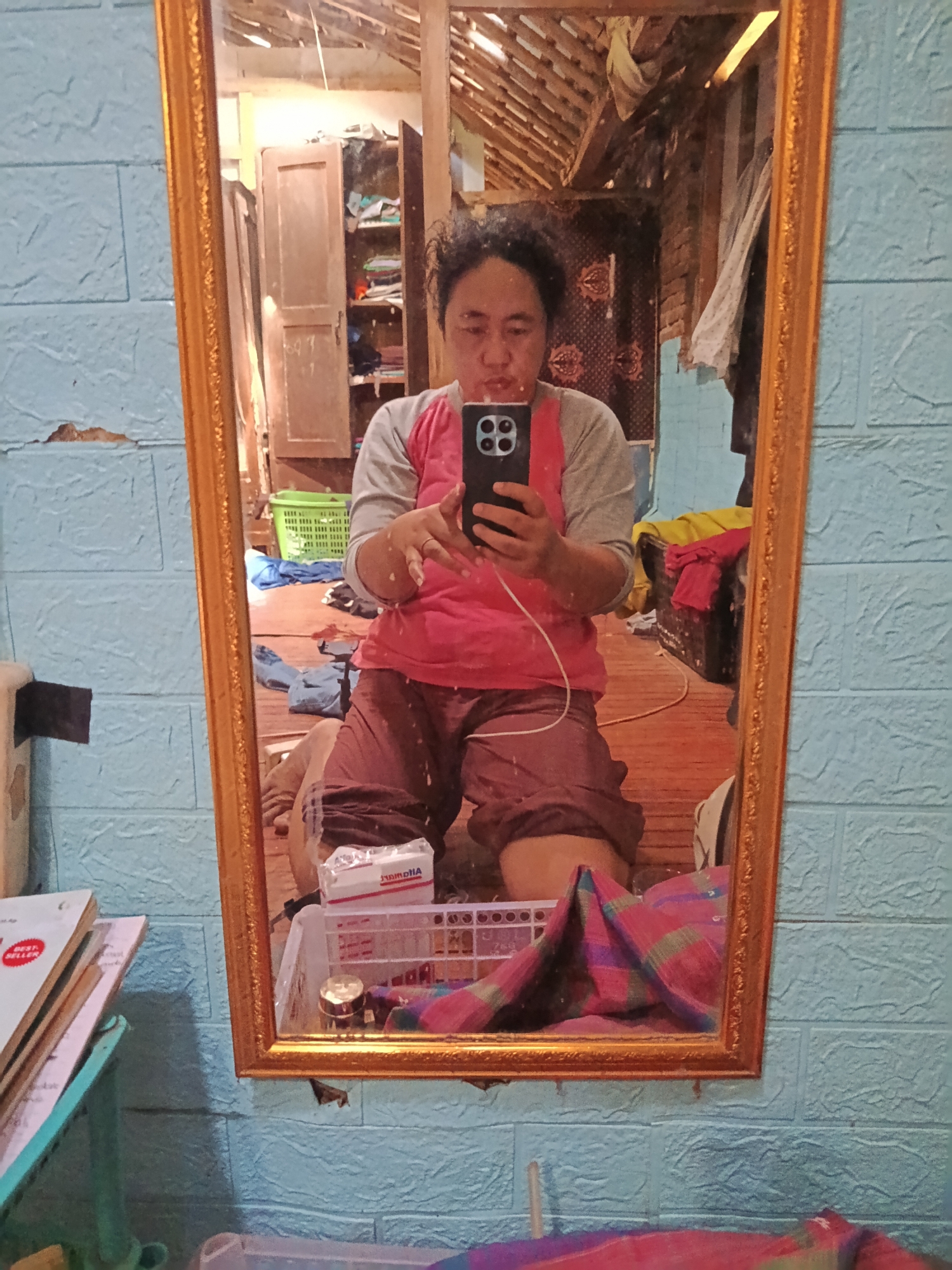 The image shows a person kneeling on a wooden floor in front of a mirror, taking a selfie. The person is wearing a pink and gray long-sleeved shirt and maroon shorts. They are holding a black smartphone in their right hand. The mirror reflects the room behind them, which appears to be a storage area with shelves, clothes, and baskets. The walls are covered with blue textured wallpaper. In the foreground, to the left of the mirror, a stack of books is visible on a teal stand. To the right of the mirror, a blanket with a red, purple, and green plaid pattern is draped over an object. The environment suggests a domestic setting, possibly a bedroom or a storage room, in Karangmojo, Indonesia, indicated by the presence of household items and typical Indonesian interior design elements.