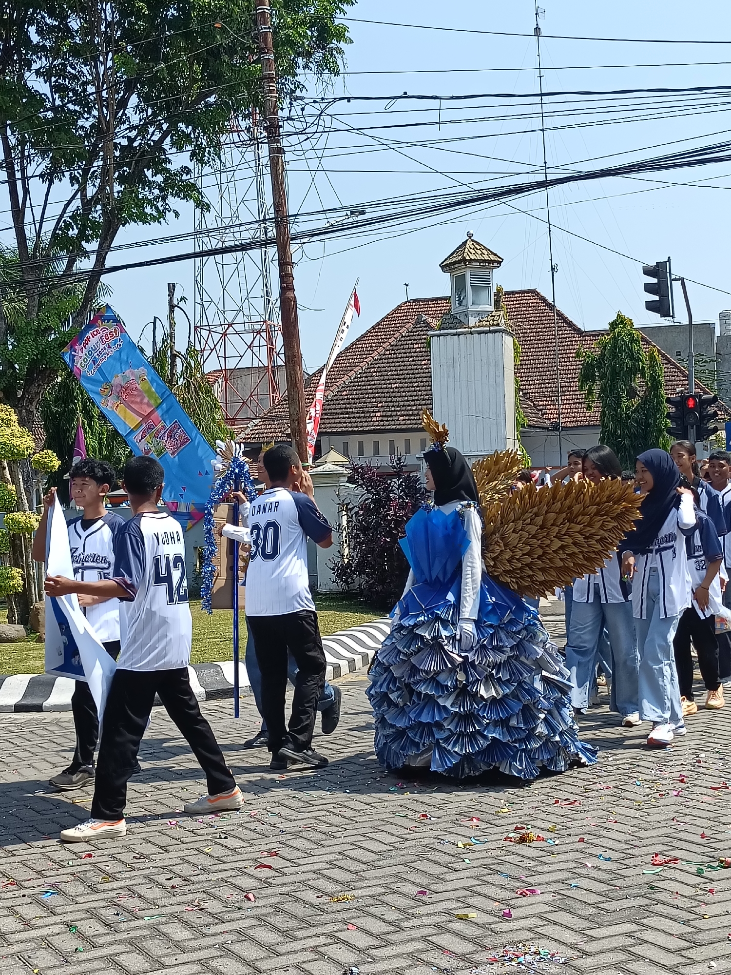 The image depicts a street parade in Kediri City, Indonesia, likely taking place on a paved road with brick-like patterns. A group of individuals, predominantly young males wearing white and blue sports jerseys with numbers and names like "YUDHA 42" and "DANAR 30" printed on them, are walking together. They are holding a large blue banner with colorful graphics and text, including "POP ICE COLOUR FEST". To the right of the group, a person is dressed in an elaborate costume featuring a large, blue, pleated dress and prominent golden wings, possibly representing an angel or a bird. The person is wearing a headscarf and gloves. Other participants, including young females in similar blue and white attire and headscarves, are also visible in the background, some smiling and interacting. The environment includes trees, power lines, a building with a tiled roof and a small tower, and a traffic light. Confetti is scattered on the ground, indicating a festive occasion.