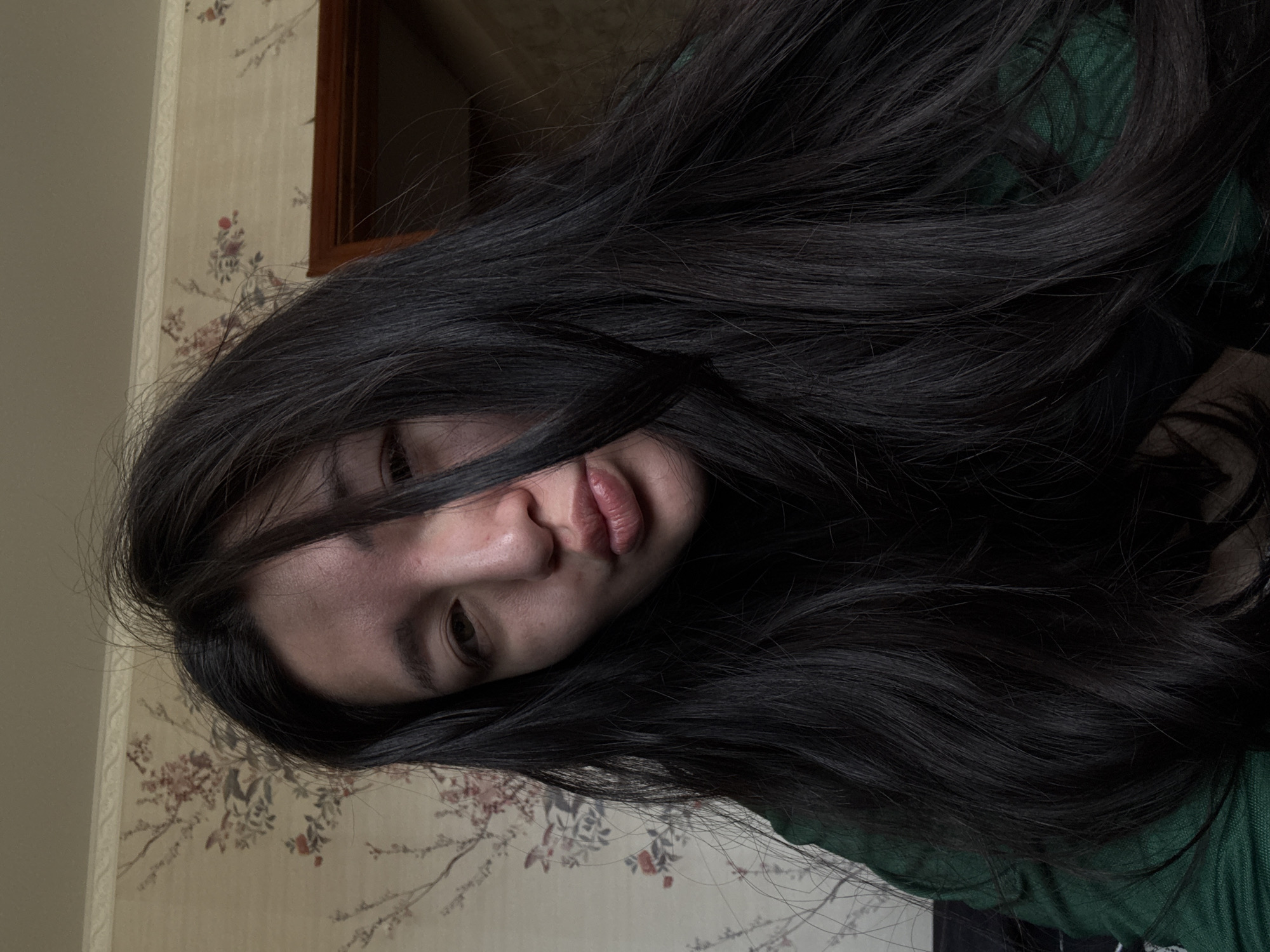 A young woman with long, dark, wavy hair that partially covers her face is positioned at an oblique angle against a wall. Her face is partially visible, with focus on her lips, nose, and one eye. She is wearing a green top. The wall behind her features a vintage wallpaper with floral patterns. A dark wooden frame is partially visible in the upper background. The photograph appears to have been taken indoors, likely in a residential setting. The precise location within Kurovskoye, Russia cannot be determined from the image.