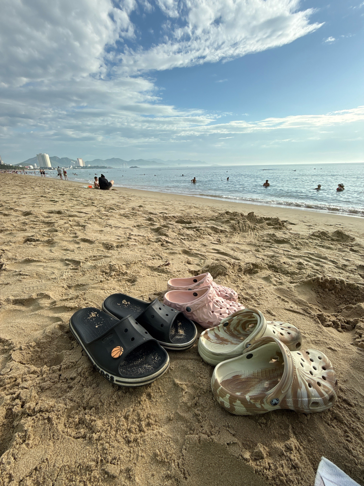 The image captures a serene beach scene in Nha Trang, Vietnam. In the foreground, several pairs of footwear are placed on the golden sand: a pair of black slides with a basketball emblem, a pair of pink clogs, and a pair of beige and brown tie-dye clogs. The sand is textured with footprints and natural undulations. In the middle ground, a gentle shoreline meets the calm sea, where a few individuals are visible wading and swimming. Further in the background, the cityscape of Nha Trang unfolds with several modern buildings and apartment complexes lining the coast, backed by rolling hills. The sky above is a mix of clear blue and scattered white clouds, illuminated by what appears to be late afternoon sunlight.