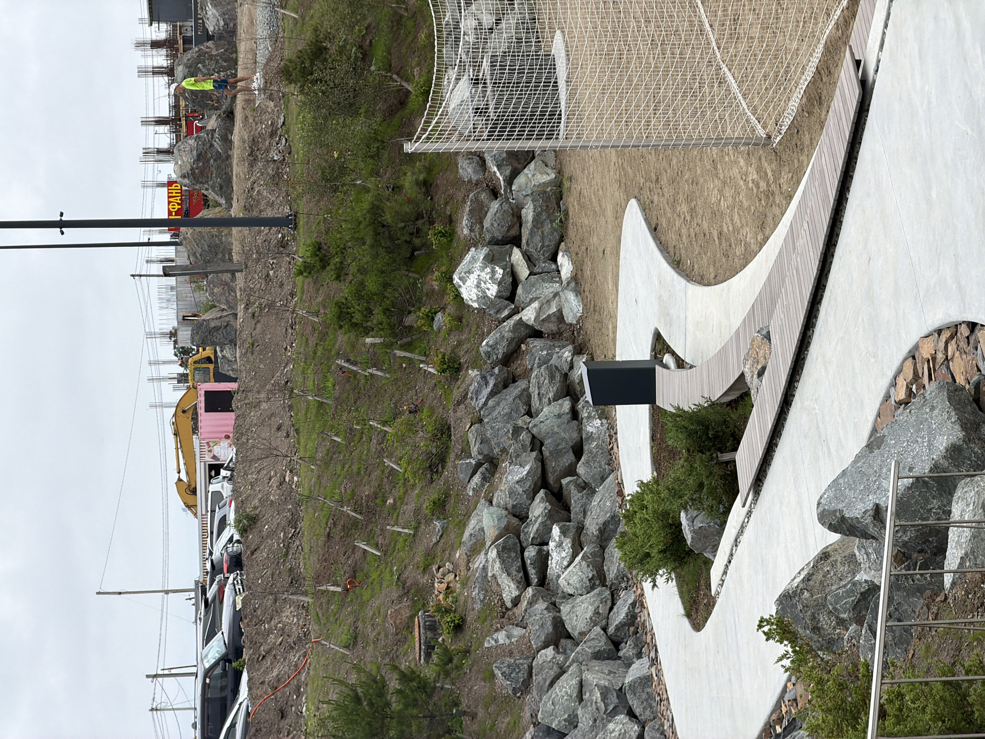 The image depicts an outdoor area with landscaping and construction. In the foreground, a curved concrete pathway winds through a rocky embankment with scattered green bushes. To the right of the pathway is a sand-covered area with a large white net, possibly part of a sports facility. In the background, a man in a neon yellow shirt and blue shorts is walking on a path. Further back, there is a construction site with a yellow excavator, large boulders, and unfinished buildings with rebar structures. A pink shipping container and several parked cars are also visible in the distance. A red sign with yellow Cyrillic lettering, reading "ФАНЬ," is mounted on a structure behind the construction. The overall scene suggests an ongoing development or renovation project in an urban or suburban setting.