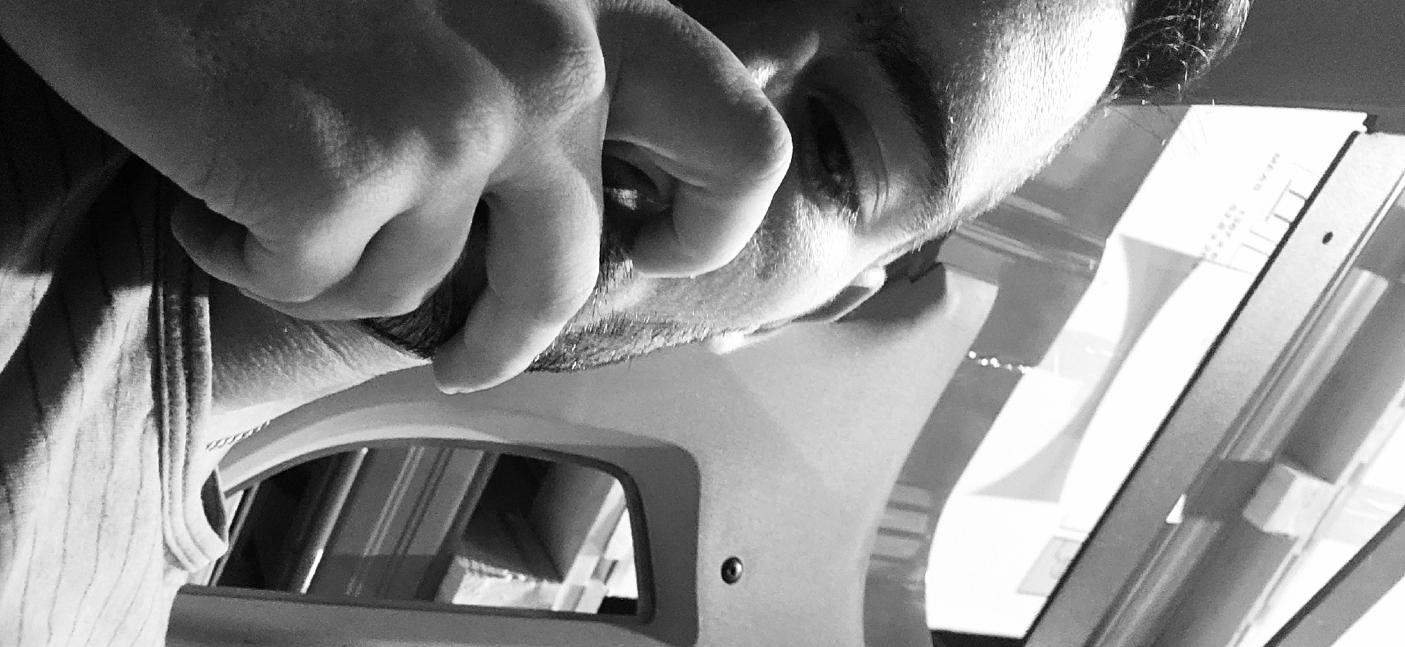 A close-up, black and white photograph depicts a person resting their hand on their face, specifically covering their eye and touching their beard. The subject appears to be looking down or off to the side. The image includes elements of a machine or device in the foreground, which appears to be reflecting an interior space. The environment is indeterminate, and there are no elements visible that would specifically place the scene in Plovdiv, Bulgaria.