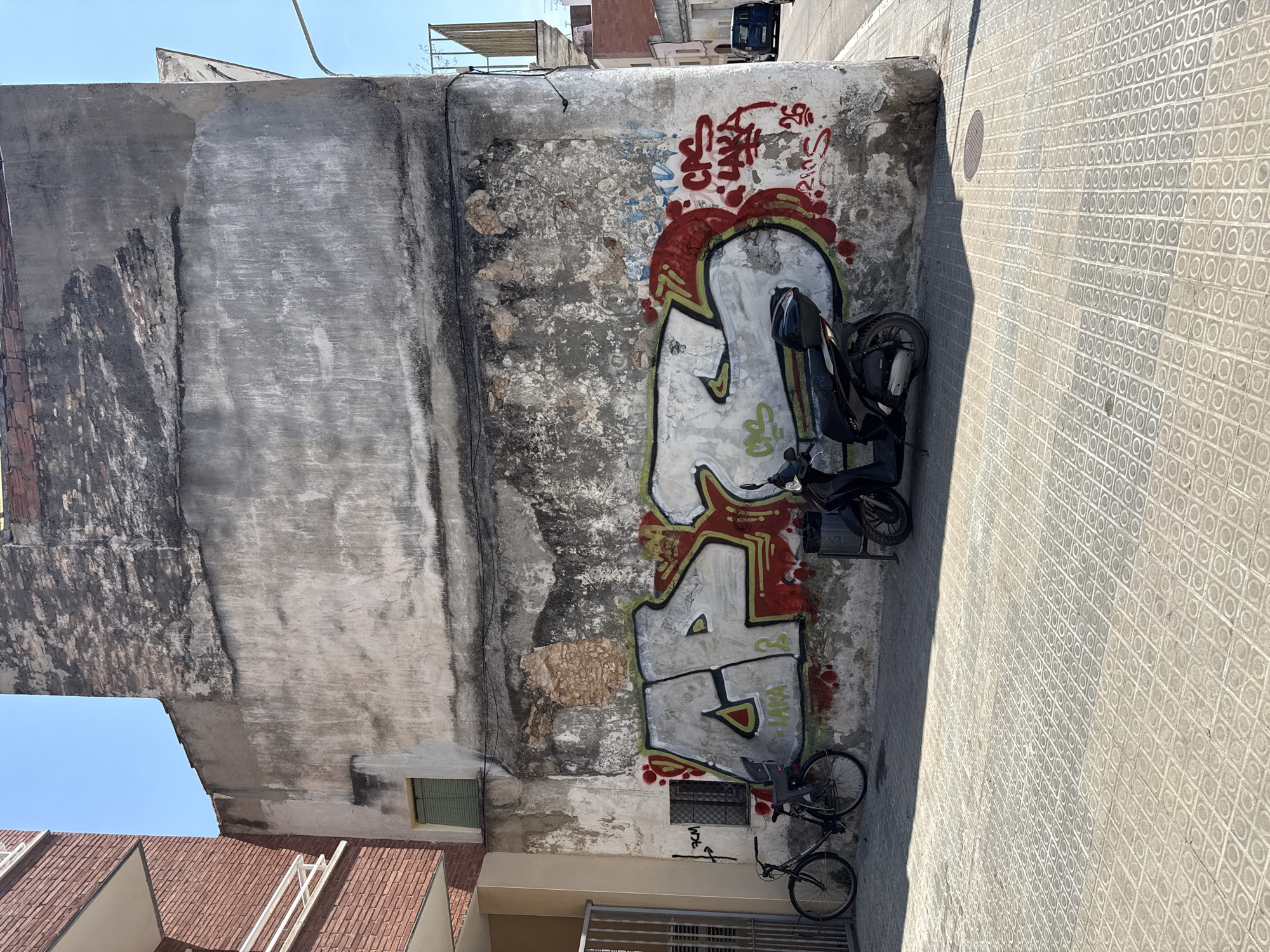 The image shows a graffiti-covered wall in an urban setting, likely Vilanova I La Geltrú, Spain. The wall is old and weathered, with patches of peeling paint and exposed brick. Large, colorful graffiti adorns the wall, featuring abstract shapes and lettering in red, white, and green. A black motorcycle is parked leaning against the wall, partially obscuring the graffiti. A bicycle is also visible, parked on the pavement nearby. The pavement is tiled with a repeating geometric pattern. The scene is bathed in bright sunlight, casting a long shadow across the pavement, suggesting it is daytime. The sky is clear and blue. The overall impression is one of an urban environment with a street art element. The graffiti on the wall appears to include the word "LAKA" in stylized lettering.