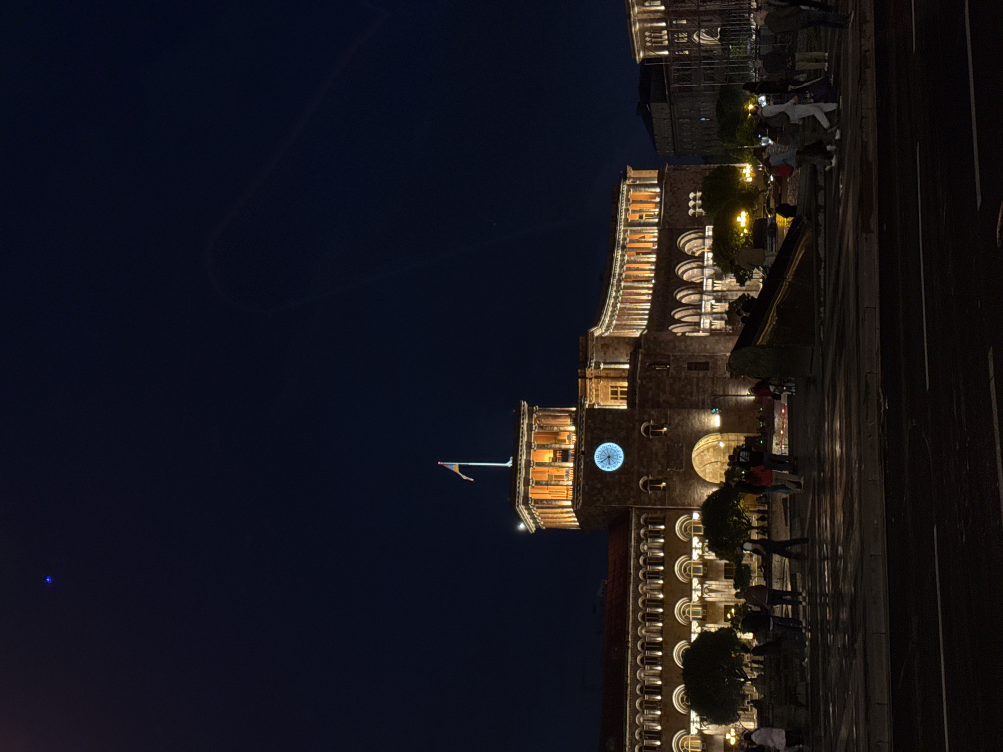 The image shows Republic Square in Yerevan, Armenia, at night. The brightly lit buildings of the square dominate the right side of the frame, and a large expanse of dark night sky occupies the left side. A flag is visible atop the central building. People are walking on the square's surface in front of the buildings.
