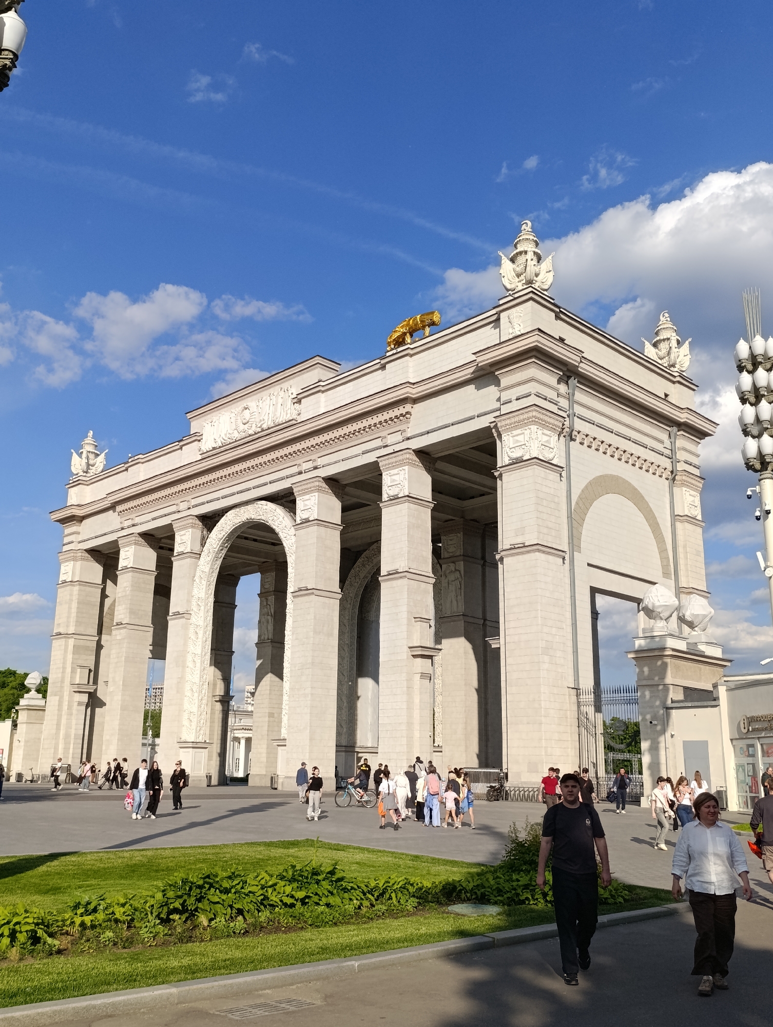The image presents a wide-angle view of a grand, pale-colored triumphal archway, likely a main entrance to a large public space in Moscow, Russia. The monumental structure exhibits a classical architectural style, characterized by multiple large arches, robust columns, pilasters, and intricate decorative friezes and reliefs.
Crowning the archway's roofline is a prominent golden sculpture of a crouching feline, possibly a lion, alongside several ornate white decorative urns or vases. A detailed relief panel is visible above the central arch. The arches are deep, with the inner surfaces of some, particularly the closest large arch, showing elaborate textural patterns.
In front of and around the archway, a paved pedestrian area is active with numerous people. Individuals and small groups are seen walking and milling about, dressed in casual contemporary clothing, indicative of leisure activity during the daytime. One person is visible riding a bicycle.
In the foreground, a neatly manicured strip of green grass and bushes provides a visual separation. The sky above is a clear, bright blue with scattered white, fluffy clouds, suggesting good weather conditions. Strong, natural sunlight illuminates the scene, casting sharp shadows.
On the far right, a tall, modern lamppost fitted with multiple light fixtures and several security cameras stands. Adjacent to it, a low-rise building features a sign with the partially visible Cyrillic text "ГИПЕР" (GIPER), suggesting a shop or hypermarket. Through the archways, glimpses of other buildings and lush greenery hint at the expansive nature of the location.