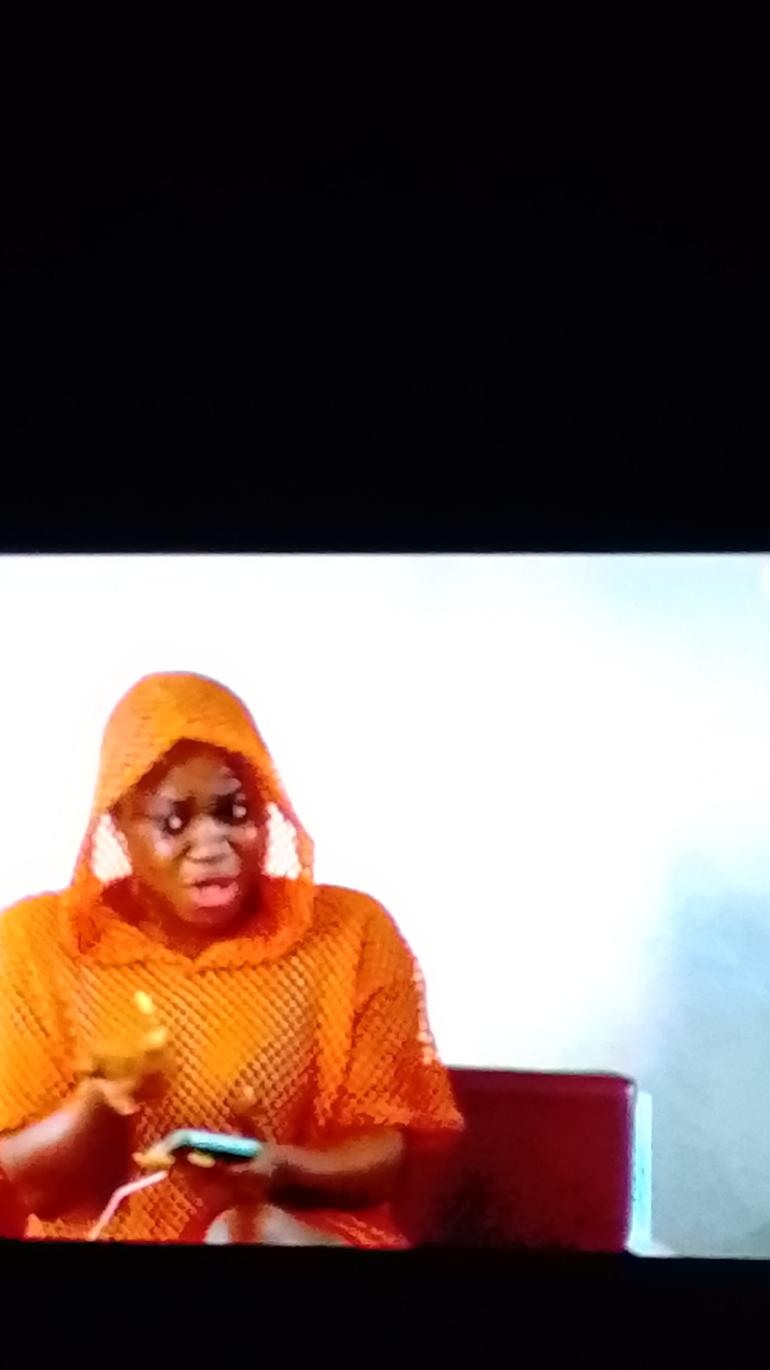 A medium shot shows a Black person, likely female, wearing an orange hooded mesh top. The person is seated, with their left hand raised and palm facing upwards, while their right hand holds a smartphone with a white cord attached. The person's mouth is open, suggesting they are speaking or reacting. The background consists of a blurred white surface on the left and a dark red chair or furniture on the right. The lighting is bright, casting a slight shadow on the right side of the person's face. The overall environment appears to be indoors, possibly a studio or a living space, in Unknown, Unknown.