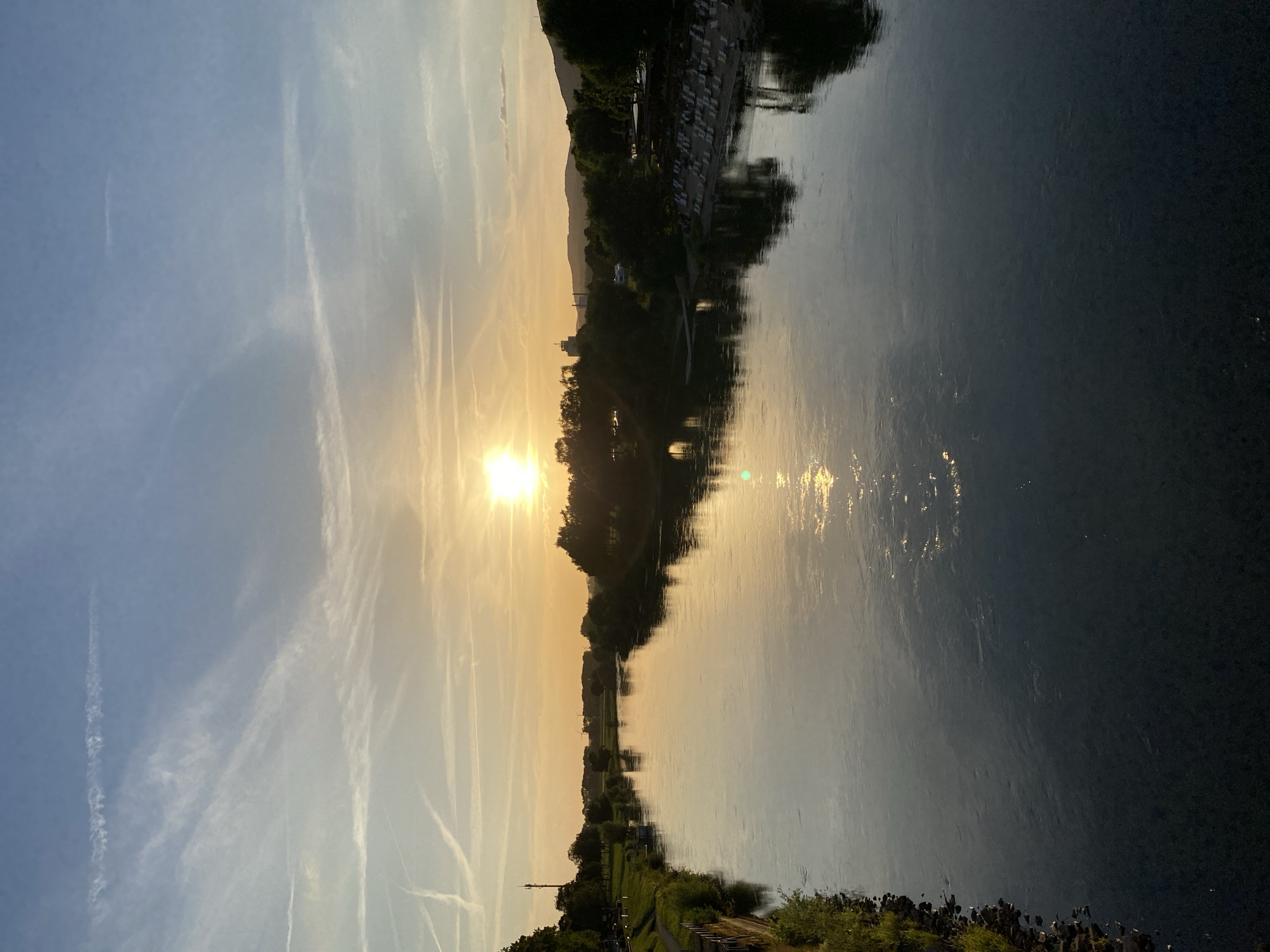 The photograph captures a serene sunset over the Weser River in Rinteln, Germany. The sky, a gradient of soft blues and warm oranges, is streaked with wispy clouds and contrails from aircraft. The sun, a bright orb low on the horizon, casts a shimmering reflection on the calm water's surface.
The riverbanks are lined with lush green trees, creating a verdant border. On the left bank, a paved walkway runs alongside a grassy area, with a few scattered trees. A narrow, dark-hulled boat is moored near the bank. On the right bank, a beach area is visible with rows of white lounge chairs arranged on the sand, suggesting a recreational space. In the distance, a cluster of trees forms a dense line, with faint outlines of hills or mountains beyond. A prominent telecommunication tower stands tall on the left bank. The overall scene is tranquil, bathed in the warm, diffused light of the setting sun.