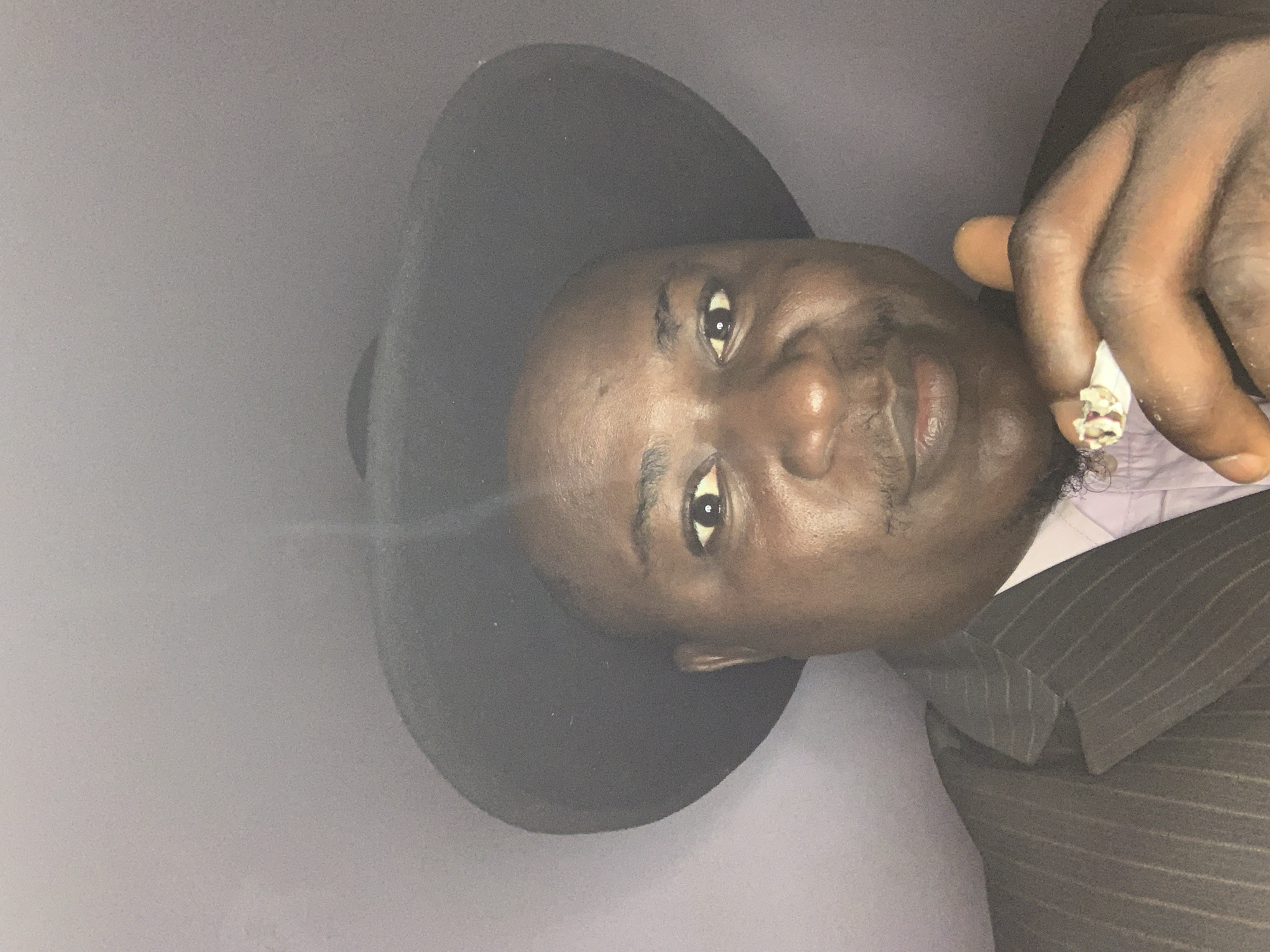 A close-up, head-and-shoulders portrait depicts a Black male adult facing directly forward, looking at the viewer. He wears a dark, wide-brimmed fedora-style hat, a dark grey or black pinstriped suit jacket, and a light lavender collared shirt. His skin is dark brown, and he has a short, dark mustache and some stubble on his chin and jawline. In his right hand, held between his index and middle fingers, is a lit cigarette with a visible ash tip, emitting a thin stream of white smoke that rises vertically. The background is a uniformly lit, plain light grey wall, suggesting an indoor setting. No specific geographical location within Akwanga, Nigeria, or any other region, is discernible from the visual information provided.