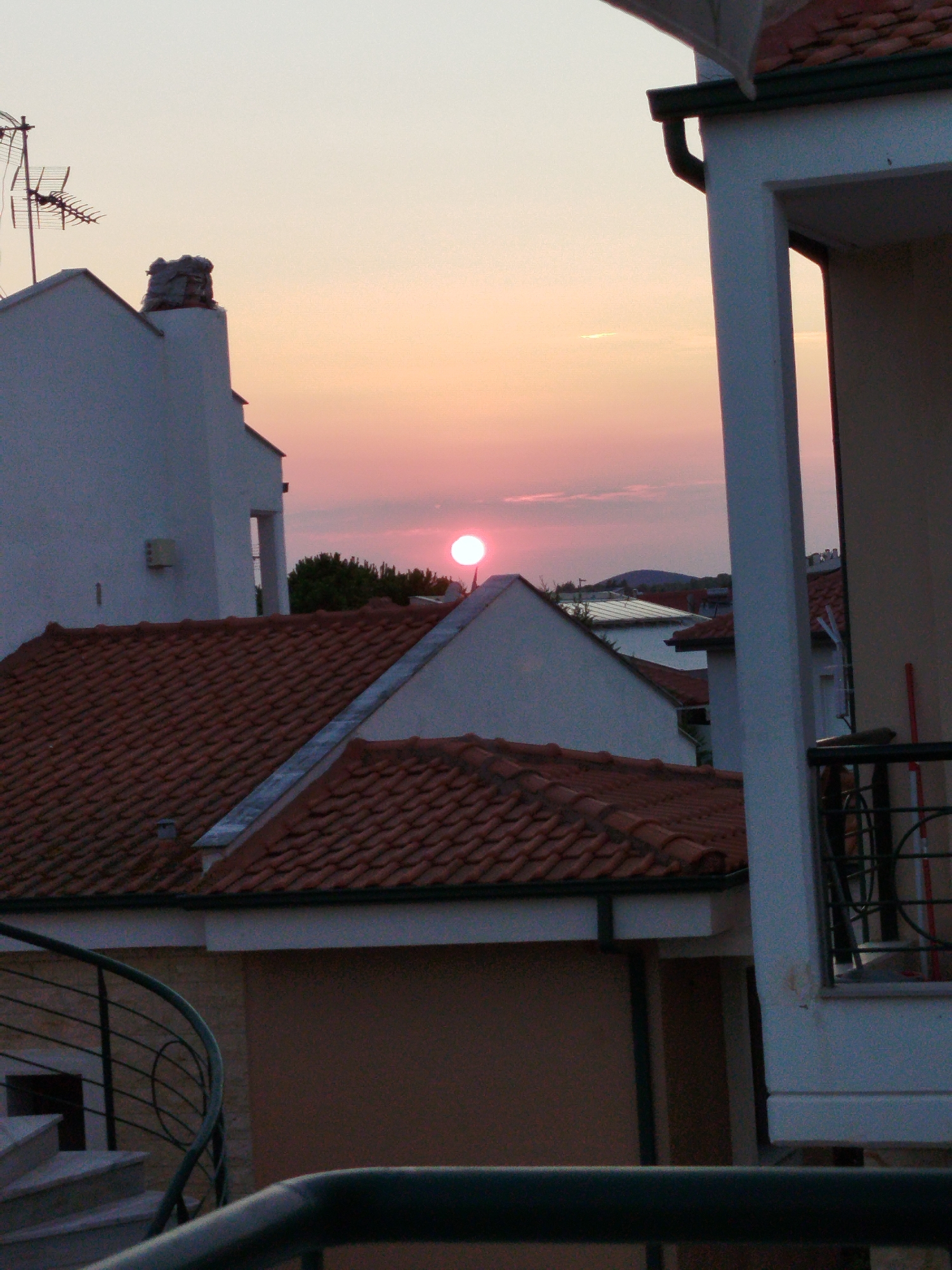 The image depicts a sunset over a residential area in Sithonia Municipal Unit, Greece. The sky is a gradient of pale orange and pink. The foreground features the roof of a building with terracotta tiles, a white wall, and a dark railing. To the left, another white building with a chimney and an antenna is visible. In the background, several other buildings with tiled roofs can be seen, and a rounded hill is present in the far distance. The sun is a bright, pink circle near the horizon. No people or animals are present in the image.