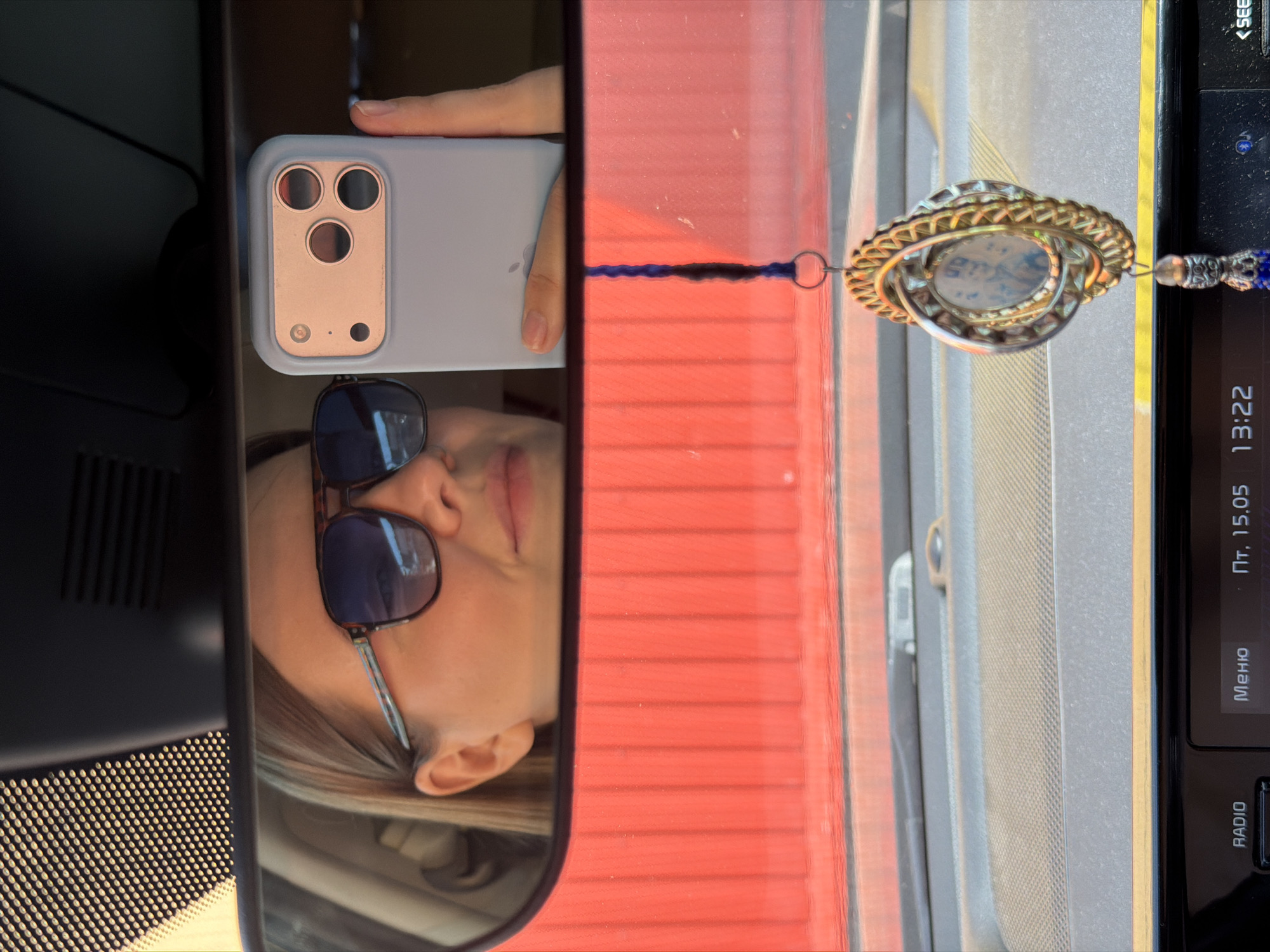 A reflective car mirror reveals a woman wearing sunglasses, holding a cell phone. The reflection includes part of the car's dashboard. An ornament hangs from the mirror. The car radio displays the date and time as Пт, 15.05 13:22. The image was taken in Chaikovsky, Russia.