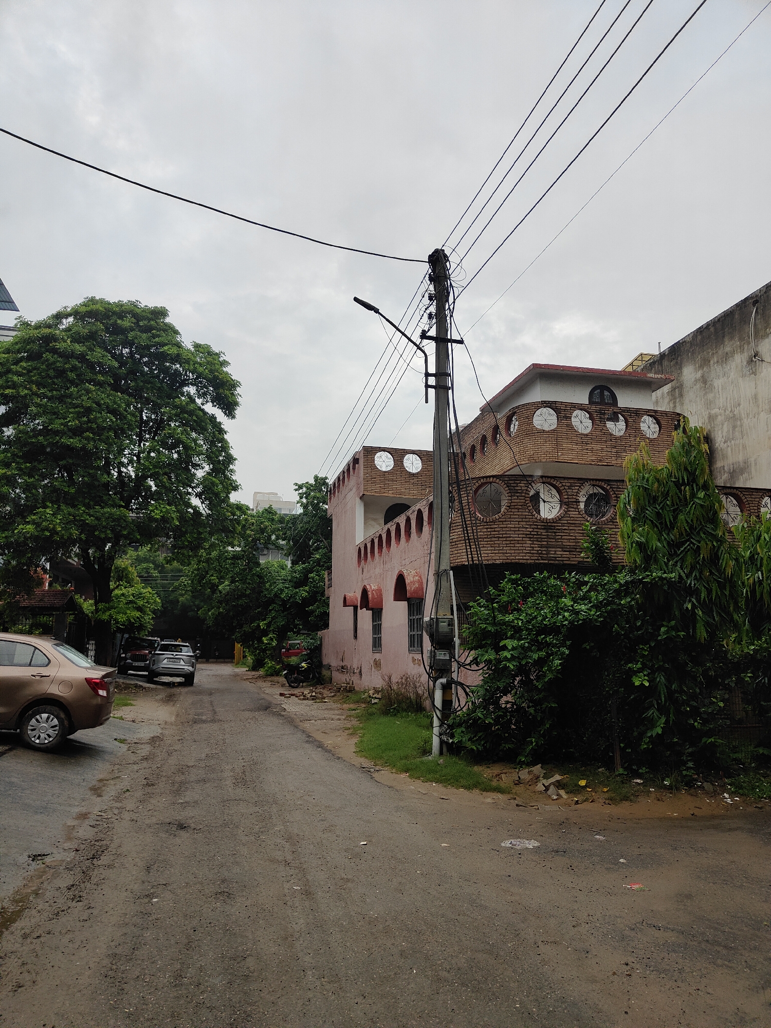 A street scene in Gurgaon, India, shows a cloudy sky overhead. A paved road, appearing worn and with some puddles, leads into the distance. On the right side of the road is a pink building with arched windows and circular decorative elements. A large tree with dense green foliage stands on the left side of the road. Several cars are parked on the left side of the street, and a motorcycle is visible on the right. A utility pole with numerous wires is prominent in the foreground on the left. The environment appears residential.