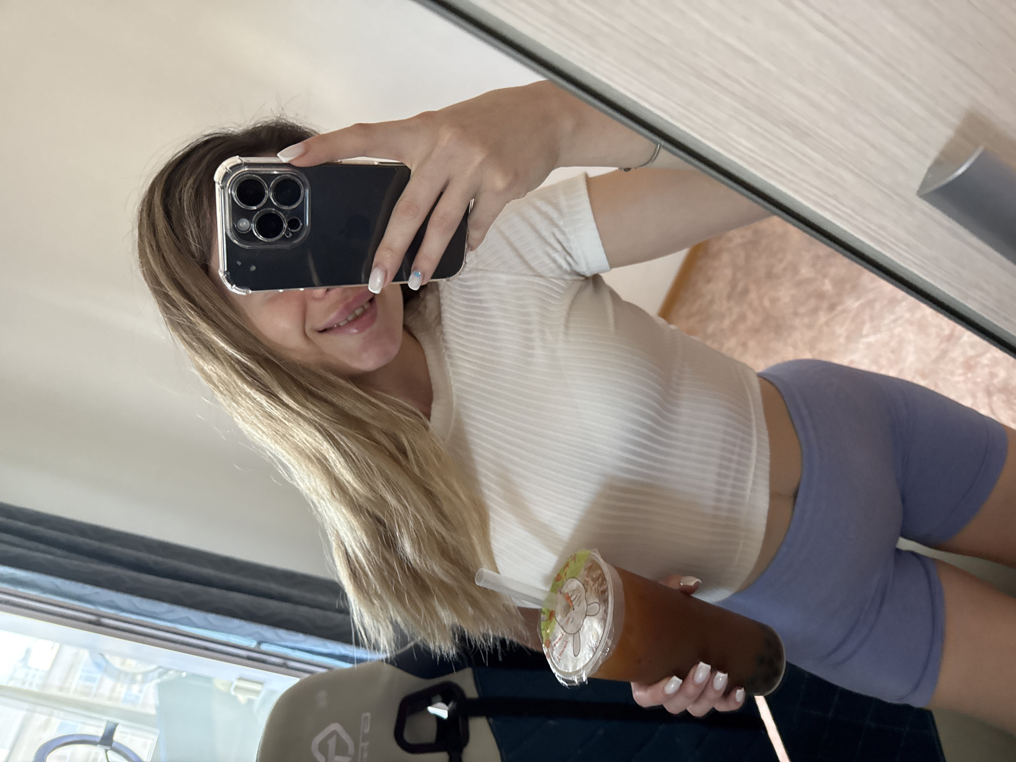 A woman is taking a selfie in front of a mirror. She is holding a black smartphone with a clear case up to her face, partially obscuring it. Her blond hair is visible on either side of the phone. She is wearing a short-sleeved white shirt and light purple athletic shorts. In her other hand, she is holding a bubble tea drink. The background shows a window with dark curtains, a gaming chair, and a partially visible rug. The location appears to be indoors, possibly in her home.