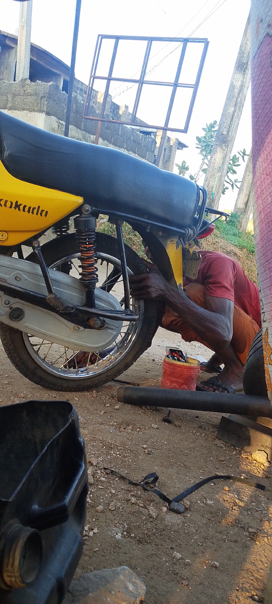 The image shows a man in Lokoja, Nigeria, working on a motorcycle tire. The motorcycle is yellow and black and has the word "Kokuda" painted on it. The man is wearing a red shirt and orange shorts and is crouched beside the motorcycle. A red bucket with items inside sits near the tire. Other objects on the ground include a black pipe and a black bucket. The background shows buildings and vegetation. The ground appears to be dirt and gravel.