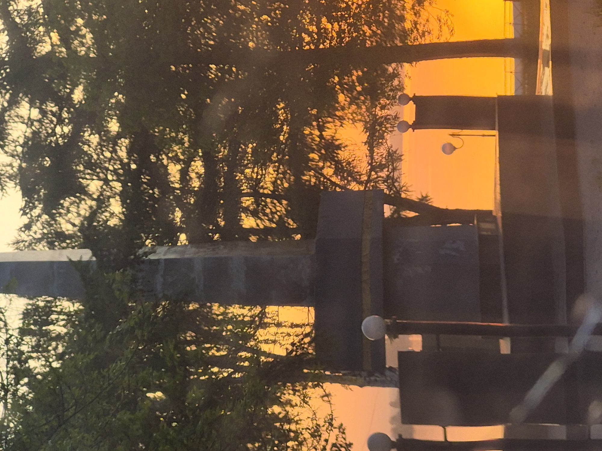 The image depicts a sunset in an urban setting. Silhouette of trees are visible in the foreground. Architectural structure is visible in the middle. Street lamps are seen on the right side of the image. The sky is orange. Location: Unknown, Unknown.
