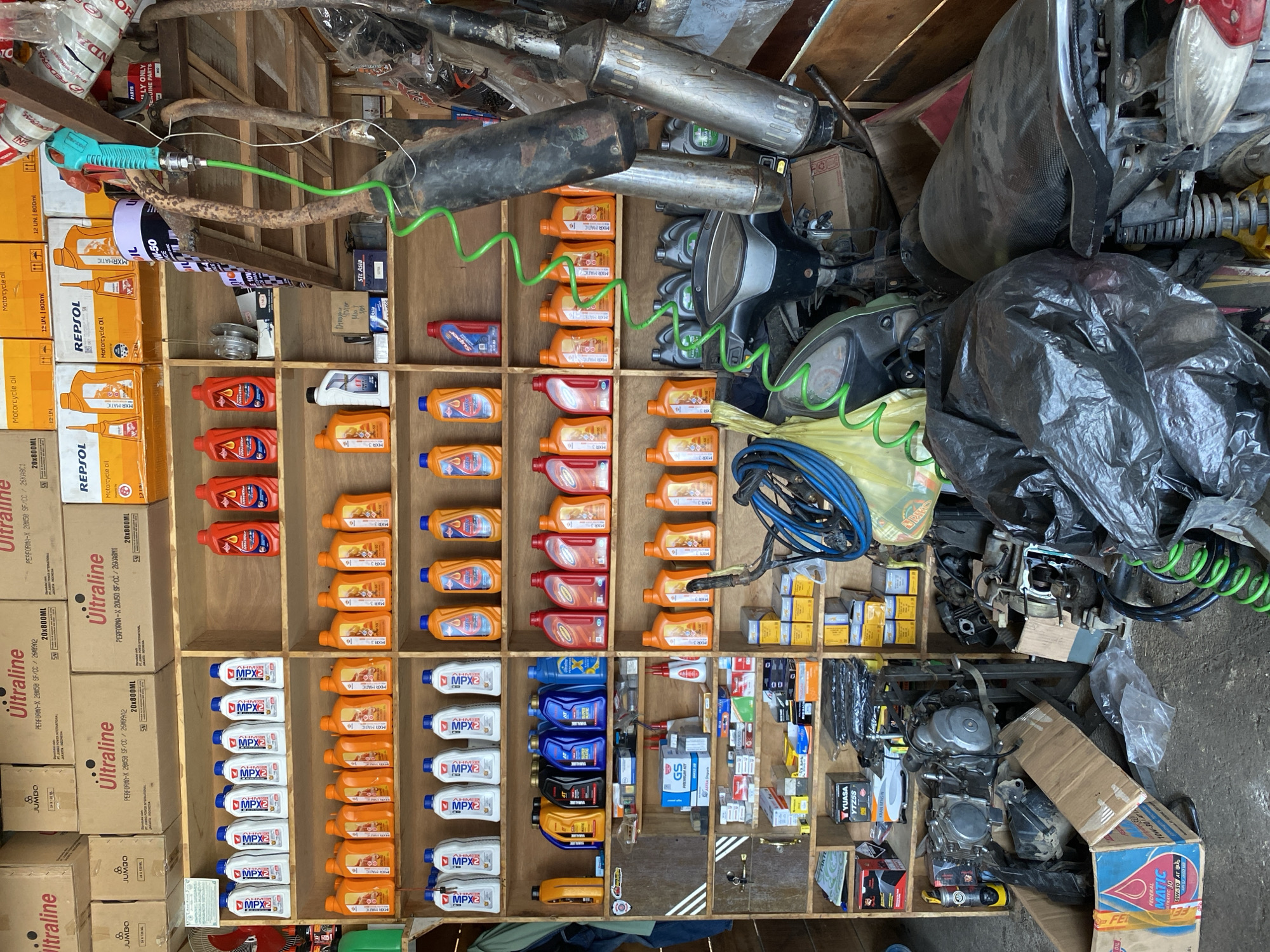 The image depicts a cluttered motorcycle parts shop, likely located in Bayu, Indonesia. Shelves are stocked with numerous bottles of motor oil and lubricants, various engine parts, and boxes of components. In the foreground, disassembled motorcycle engines and body parts are visible, some covered in black plastic bags. A green hose, possibly for an air compressor, snakes across the scene, connecting to a nozzle hanging from the ceiling. The overall impression is one of a busy, well-stocked, but somewhat disorganized repair shop.