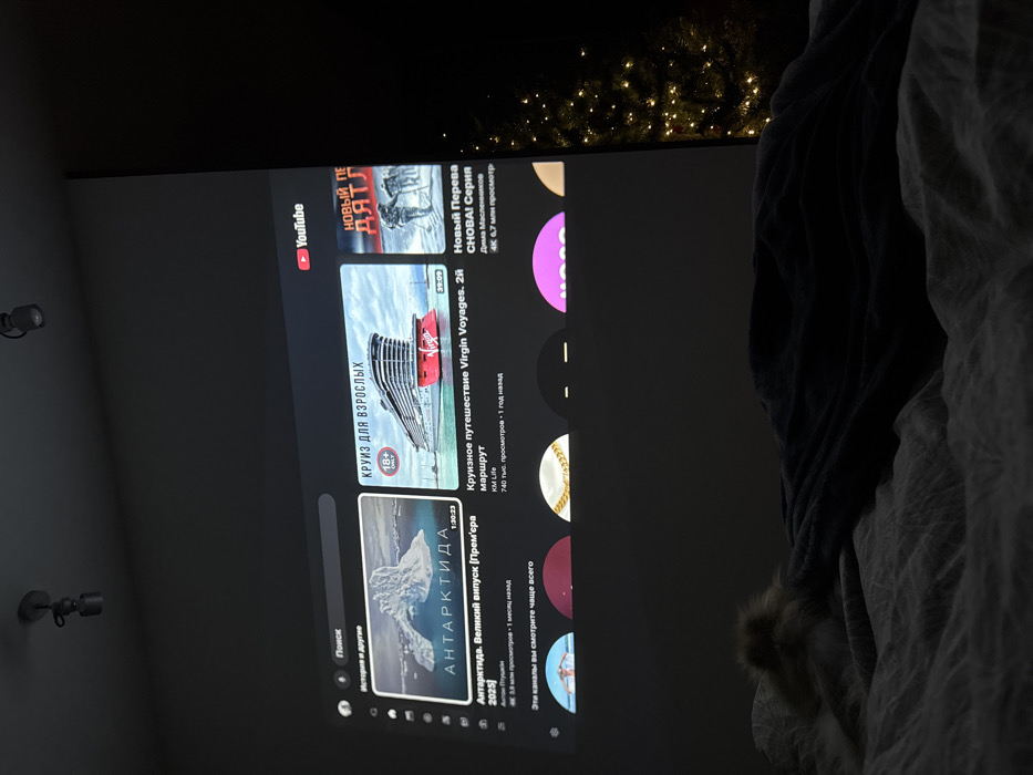 The screen shows the YouTube interface with Russian text. A YouTube logo is in the top left corner. The screen displays recommended videos, including a "Cruise for Adults" featuring a Virgin Voyages ship, and "Antarctica" by Anton Ptushkin. It also displays "New Series Again!", "Cruise travel Virgin Voyages". There are thumbnails of other videos and channels. The interface is slightly tilted.