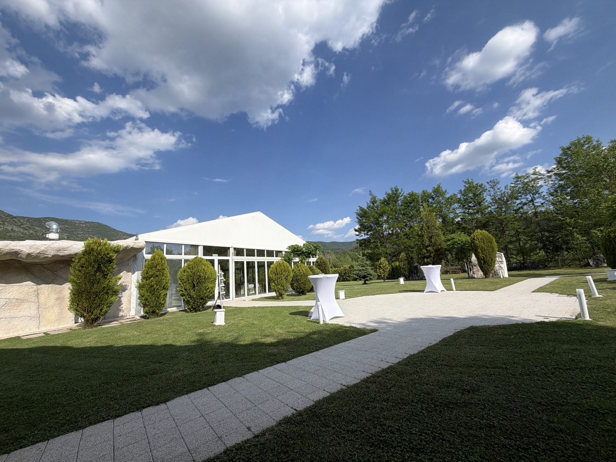 This forensic description details an outdoor event venue in Koprivlen, Bulgaria, under a clear blue sky with scattered white clouds. The central feature is a large white tent-like structure with a peaked roof and a glass facade, revealing a glimpse of an interior space. The structure is flanked by neatly trimmed conical evergreen shrubs and, on its left, a textured light-colored stone-clad wall with a prominent metallic vent or chimney pipe on top.
A light-colored paved pathway, possibly made of small pavers or gravel, traverses the well-maintained dark green lawn, forming a junction in the midground. Two white high-top cocktail tables, covered with fitted white fabric, are positioned at this pathway intersection. Several white cylindrical bollards or lights are spaced along the pathway and within the lawn area.
The background features verdant hills covered with vegetation, while a variety of green trees and additional shrubs line the perimeter of the venue, separating it from the distant natural landscape. The scene is devoid of people or active events, suggesting a prepared or unoccupied state. Sunlight is evident, casting subtle shadows across the lawn and pathway.