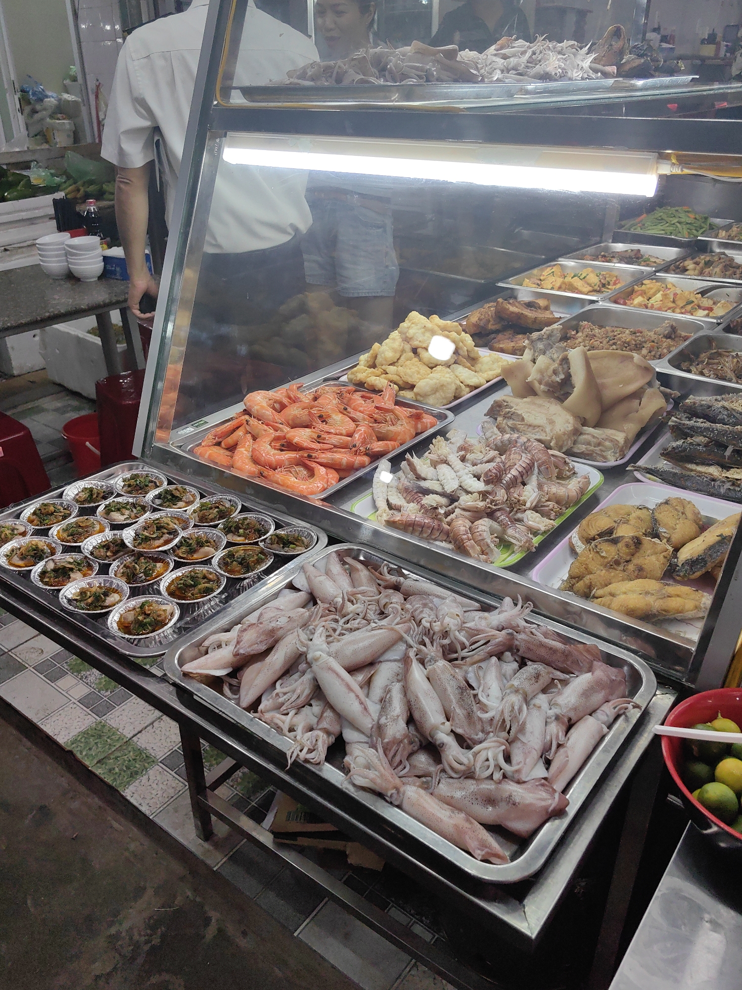 A street food vendor display in Hải Phòng, Vietnam, features a variety of seafood and prepared dishes under a glass-fronted, illuminated display case. The trays contain raw squid, cooked shrimp, fried fish, and an assortment of small foil dishes with visible herbs and spices. A red plastic bucket containing limes and a white plastic spoon sits to the side. A man in a white shirt stands behind the display case, presumably a vendor, while another person is partially visible in the background. The scene suggests a local eatery or market setting.