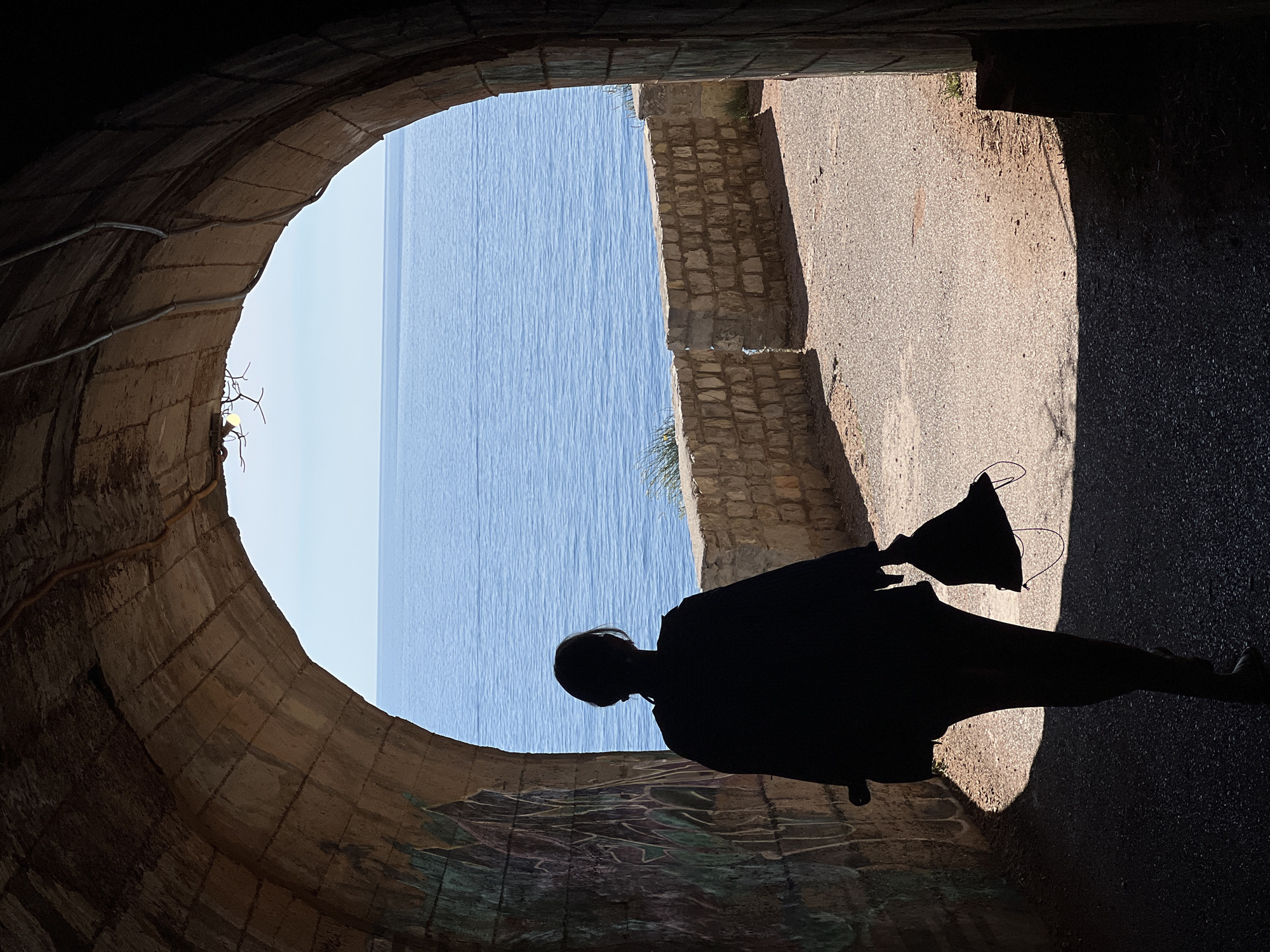 A single individual, presented as a full-body silhouette, walks away from the viewer, emerging from a dark, arched stone tunnel towards the bright daylight and the sea in Perazića Do, Montenegro. The tunnel is constructed from large, rough-hewn stone blocks, forming a distinct archway. The interior of the tunnel exhibits visible graffiti on the left wall, featuring abstract shapes in shades of green and purple. Along the upper curve of the arch on the right, several light-colored pipes or conduits are affixed, and near the top left, a bare light bulb with associated wiring or structural elements emits a faint yellow glow.
The person, of indeterminate gender, is dressed in dark, loose-fitting attire, and carries a dark, soft bag with visible straps in their right hand. Their silhouette is starkly contrasted against the bright exit of the tunnel.
Outside the tunnel, a light-colored, reddish-brown gravel path is illuminated by sunlight. To the right of the path, a low, weathered stone wall, constructed from rectangular blocks, extends towards the background, with a noticeable break or opening in its structure further along. Beyond the wall and path, a vast expanse of calm, deep blue sea extends to a clear horizon under a lighter blue sky. Sparse green vegetation is visible on the terrain immediately outside the tunnel and behind the stone wall. Strong shadows from the tunnel archway are cast upon the initial section of the sunlit path, marking the transition from the dark interior to the illuminated exterior.
