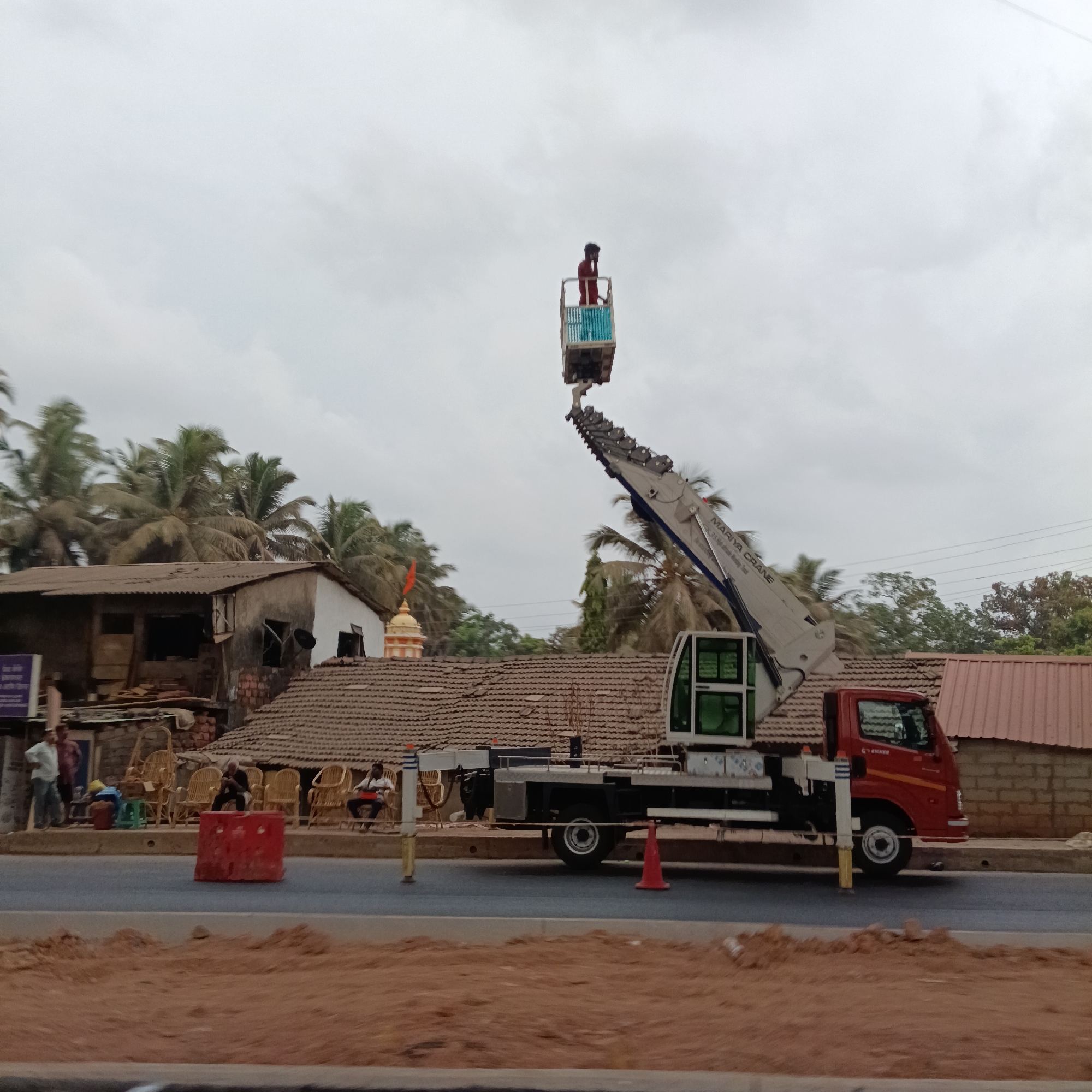 The image shows a man standing in a crane bucket high in the air in Sangolda, India. Below the man, a red Eicher truck with a white Mariya Crane boom is parked on the road. Several people are seated along the roadside near a red traffic barrier. A cluster of buildings with terracotta tile roofs are behind the truck. Palm trees are visible in the background under a cloudy sky. A Hindu temple dome with an orange flag is also in the background. A road under construction is in the foreground.