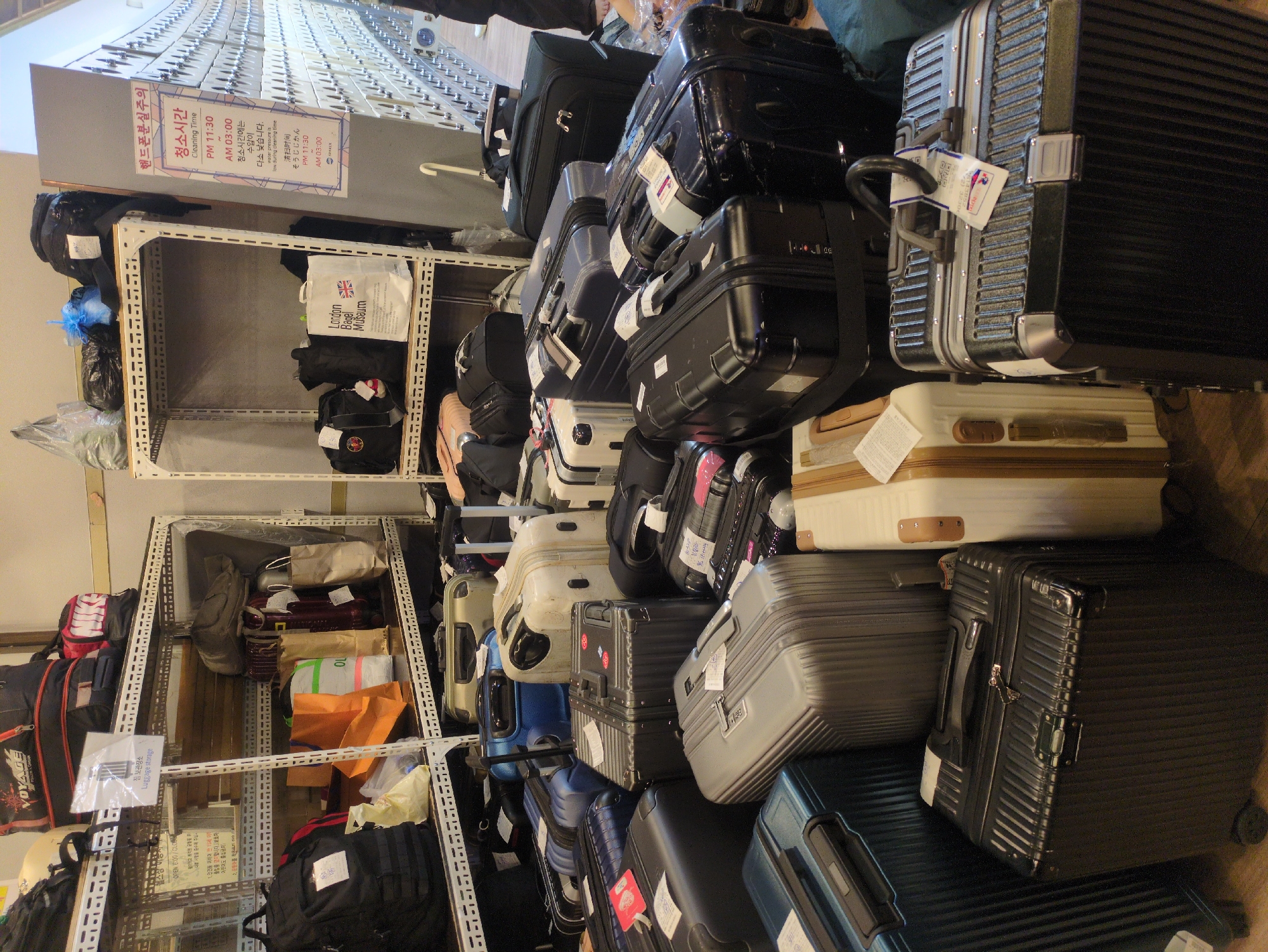 The image depicts a luggage storage area. Various suitcases and bags of different sizes and colors are stacked on shelves and piled on the floor. A sign in Korean and English warns about cleaning times and reduced water pressure. There are shopping bags from "London Bagel Museum." There is also a sign "Luggage Storage." This is likely taken in Seoul, South Korea, given the text in Korean.