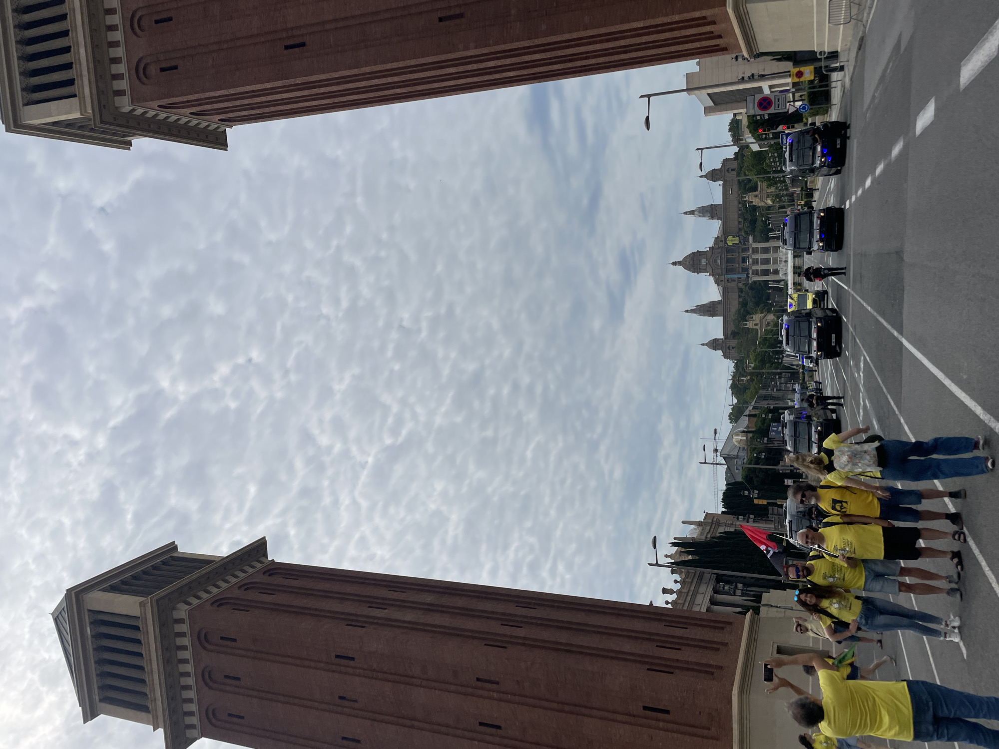 The image depicts a view from a street in Barcelona, Spain, framed by two tall, brick towers. In the distance, a grand building with multiple domes and towers, likely the Palau Nacional on Montjuïc, is visible atop a hill. A procession of dark-colored police vans and an ambulance is moving down the street towards the camera. Several people in yellow t-shirts are standing on the street, some posing for a photograph, while others are walking away. One person on the left is holding up a phone to take a picture. The sky is overcast with scattered clouds. The overall scene suggests a public event or demonstration in an urban setting.