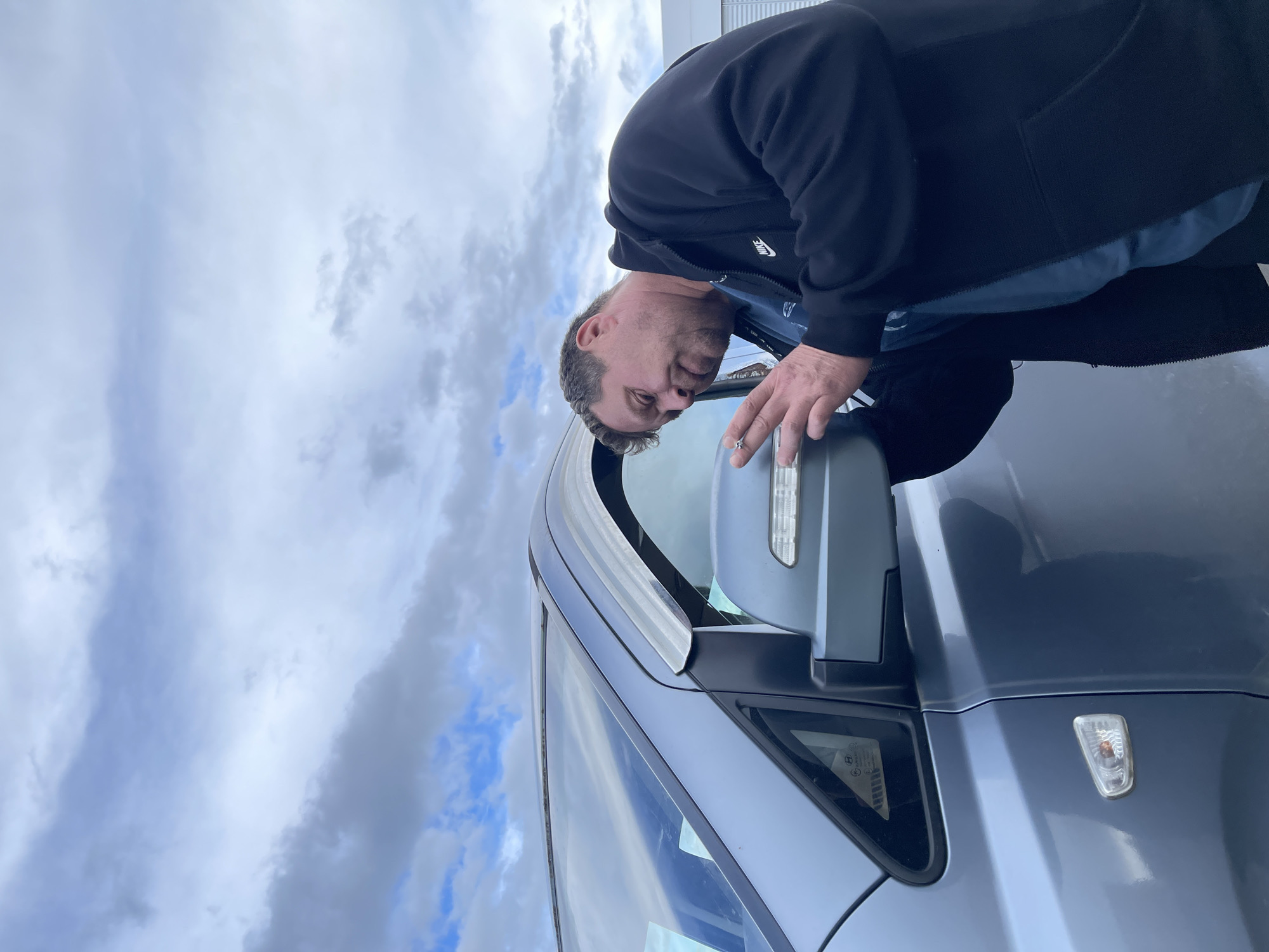 A man is standing outside near a gray car. He is wearing a black hooded jacket and appears to be examining the car's side mirror. The sky is cloudy. The location is likely in a parking area in Бавыкино, Russia, although this cannot be definitively confirmed from the image.