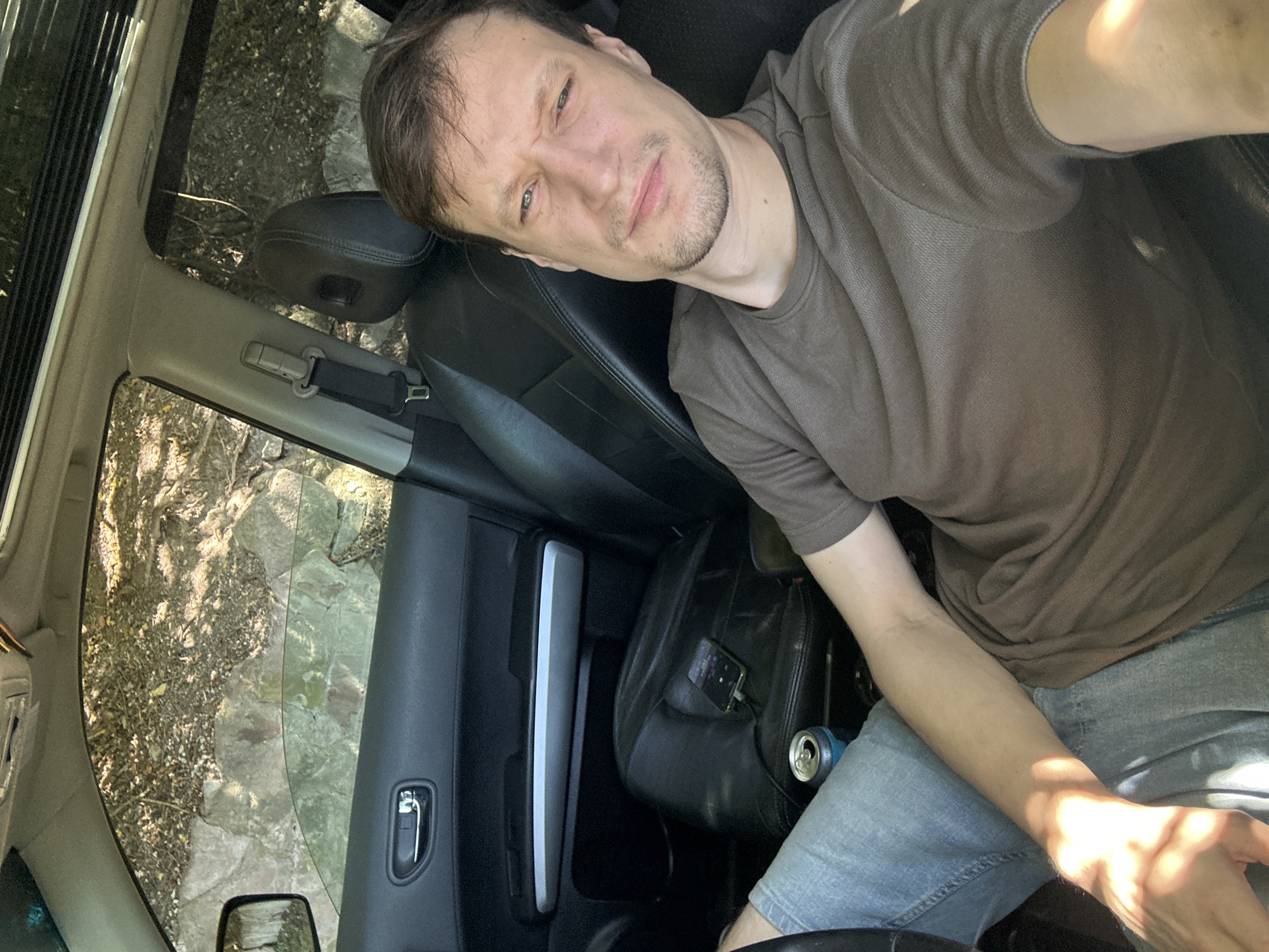In the original image, a man is sitting in the driver's seat of a car. He is wearing a brown t-shirt and blue jeans. He is looking at the camera with a strained expression. A can of soda is visible in the center console of the car, and a phone is plugged into a charger on the passenger seat. The interior of the car appears to be black leather. The windows of the car are open, and the outside environment is visible. The outside environment appears to be a wooded area with rocks and trees. The image was taken in Makiv, Ukraine.