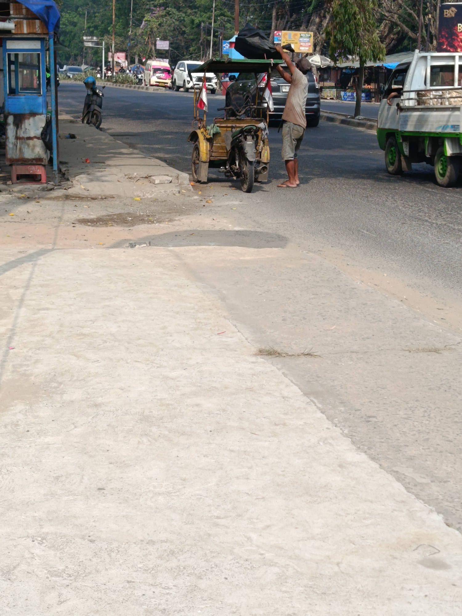 A man stands on the right side of a street in Palembang, Indonesia, holding a black tarp. He is wearing a light brown t-shirt and grey shorts. Next to him is a three-wheeled vehicle with a yellow body and a green canopy. In the background, a white truck with a green chassis is parked. The street is paved with asphalt and has a concrete sidewalk on the left side. Several vehicles are visible on the road, including cars and motorcycles. A small blue booth is located on the left side of the sidewalk. The scene appears to be a street with a mix of commercial activity and transportation.