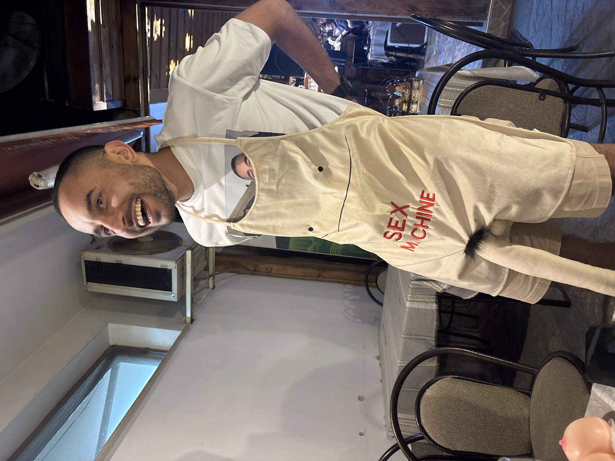 An adult male is standing in what appears to be a restaurant or bar. He is wearing a white t-shirt with an image of another male's face on it and a light-colored apron that resembles a phallus with "SEX MACHINE" embroidered in red letters. The apron has two black dots for eyes and a drawn mouth. A furry object is attached to the lower part of the apron, extending downwards and resembling male genitalia. The man is laughing and looking to his left. Behind him, tables are set with tablecloths and chairs. In the background, there are wooden beams and a glimpse of other patrons. The overall setting suggests a casual and possibly humorous environment. The provided information states the location is the city of Karlovo, Bulgaria, though this cannot be independently verified from the image alone.