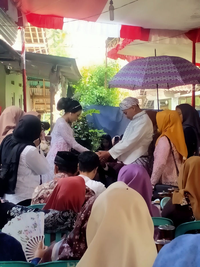 A wedding ceremony takes place under a tent with red and white draping. The bride and groom, dressed in traditional attire, are holding hands. Guests are seated in chairs facing the couple. Many women are wearing headscarves, including shades of yellow, purple, and beige. One person is holding a white fan. An ornate purple umbrella provides shade to the couple. Trees and buildings are visible in the background.