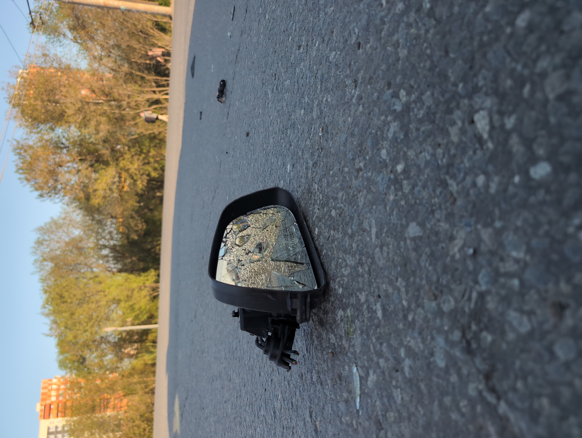 On a paved road, a car side mirror with shattered glass lies on its side. The mirror housing is black. Part of the mounting mechanism and wires are visible. The background shows trees, a building with orange accents, and a light blue sky. This location is in Izhevsk, Russia.