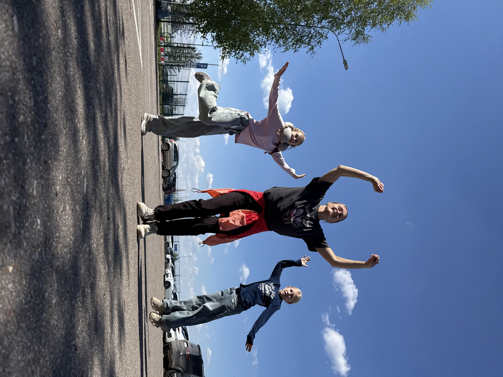 Three people are posing with outstretched arms in a parking lot at Аэропорт Уфа, Russia. The parking lot contains parked cars. The sky is blue with some clouds. There is a tree and a light post.