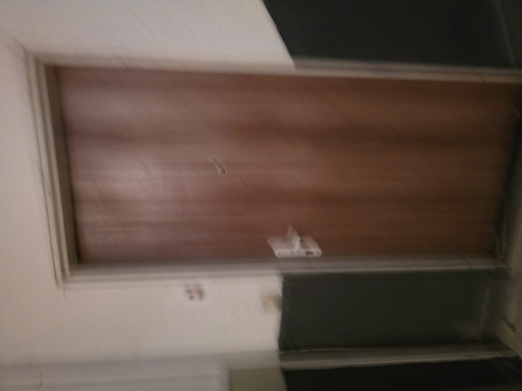The image is a slightly blurry, medium-angle shot of a closed wooden door in an interior hallway. The door is a medium brown wood grain and has a simple silver door handle and a small, circular peephole. To the left of the door, a portion of a white wall is visible, with a small rectangular object with red and blue markings attached to it. Below the white wall and to the left of the door, there is a dark grey or black wainscoting. To the right of the door, the wall is white and then transitions to dark grey wainscoting. The floor appears to be a light colored tile. The lighting is dim, creating a somewhat shadowed effect on the lower portions of the scene. The image is a photograph of a door in an apartment building or similar residential setting. The context of Yambol, Bulgaria, is not visually ascertainable from the image itself.