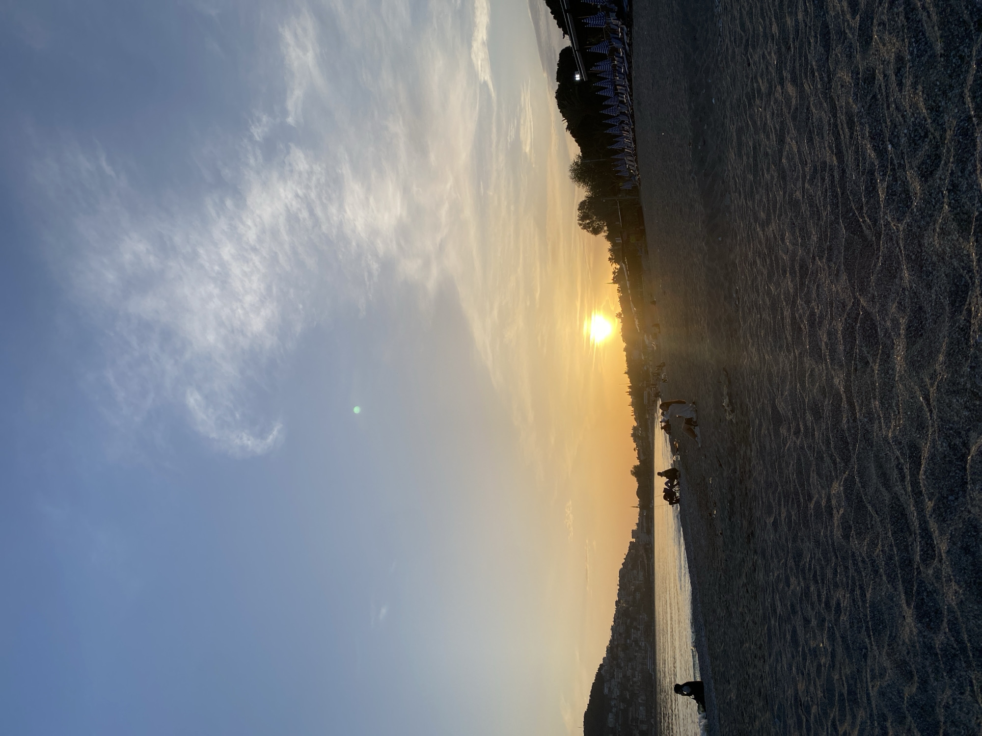 The image depicts a coastal scene at sunset in Alanya, Turkey. The sun is setting over the water, creating a golden glow in the sky. A beach with sunbeds and trees lines the upper right of the frame. People are scattered along the beach near the waterline. The ocean gently meets the shoreline. The sky transitions from a clear blue to a golden hue, with wispy clouds. A cityscape stretches along the coast.