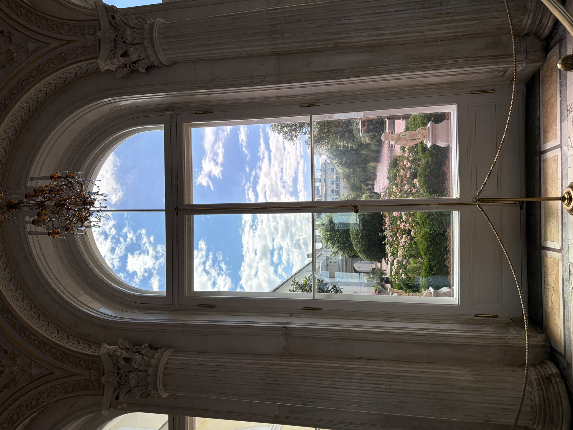 The image shows a view through a large, ornate arched window with a central rectangular pane, looking out onto a beautiful garden on a bright, sunny day. The sky is a vibrant blue filled with fluffy white clouds. The garden appears manicured, with lush green bushes and abundant pink roses in bloom. A classical statue of a nude figure stands on a pedestal in the garden, adding to the timeless elegance of the scene. In the background, a light-colored building with a mansard roof is visible, suggesting a historic or grand estate.
Inside the room, the window frame is made of white wood and is divided by a horizontal mullion. The interior architecture is grand and classical, with detailed moldings and a large, elaborate chandelier with crystal elements hanging in the upper left corner, partially obscured by the window. The walls appear to be made of marble or stone, with decorative columns and intricate plasterwork visible on the left side of the frame. The overall impression is one of refined beauty and tranquility, with the garden bathed in sunlight creating a peaceful atmosphere. The time of day appears to be midday, given the bright sunlight and clear sky. The location is Saint Petersburg, Russia, which is evident from the architectural style and the general ambiance associated with the city's palaces and gardens. There is no visible text in the image.