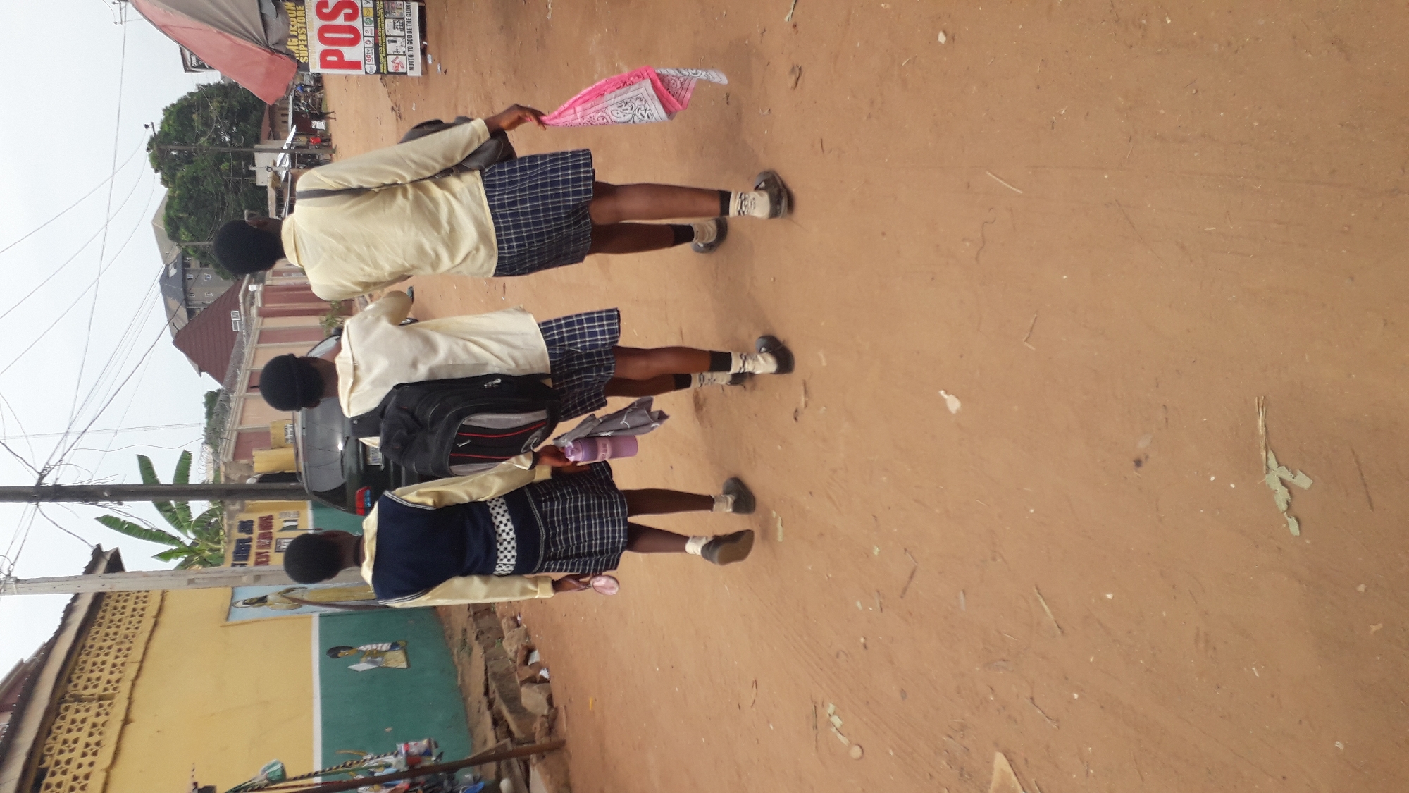 Three schoolgirls are walking on a brown dirt road in Onitsha, Nigeria. They are all wearing school uniforms consisting of a light-colored shirt and a blue and white plaid skirt. The girl in front is holding a pink and white patterned cloth. One of the girls is carrying a backpack. The road is lined with buildings, including a yellow building with a mural and a store with a red and white awning. Overhead power lines are visible.