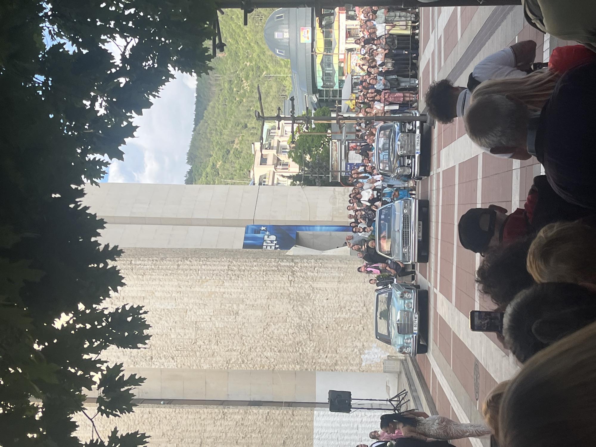 The image shows a public event in Blagoevgrad, Bulgaria, likely a motorcade. Three vintage cars are prominently displayed on a red and white tiled street, lined with crowds of people. In the background is a building with a distinctive dome and green hills. The foreground is partially obstructed by trees and people, some of whom are recording the event with their phones. A building with a light stone facade is visible to the left. A speaker setup suggests an official event.