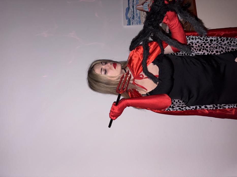 A person with blonde hair wearing a black dress, red gloves, and a red cape with black fur trim poses for a photo. They hold a red trident in one hand and have a large black spider perched on their shoulder. A patterned shawl is draped over them. A picture hangs on the wall in the background, and a pale wall takes up most of the left side of the image.