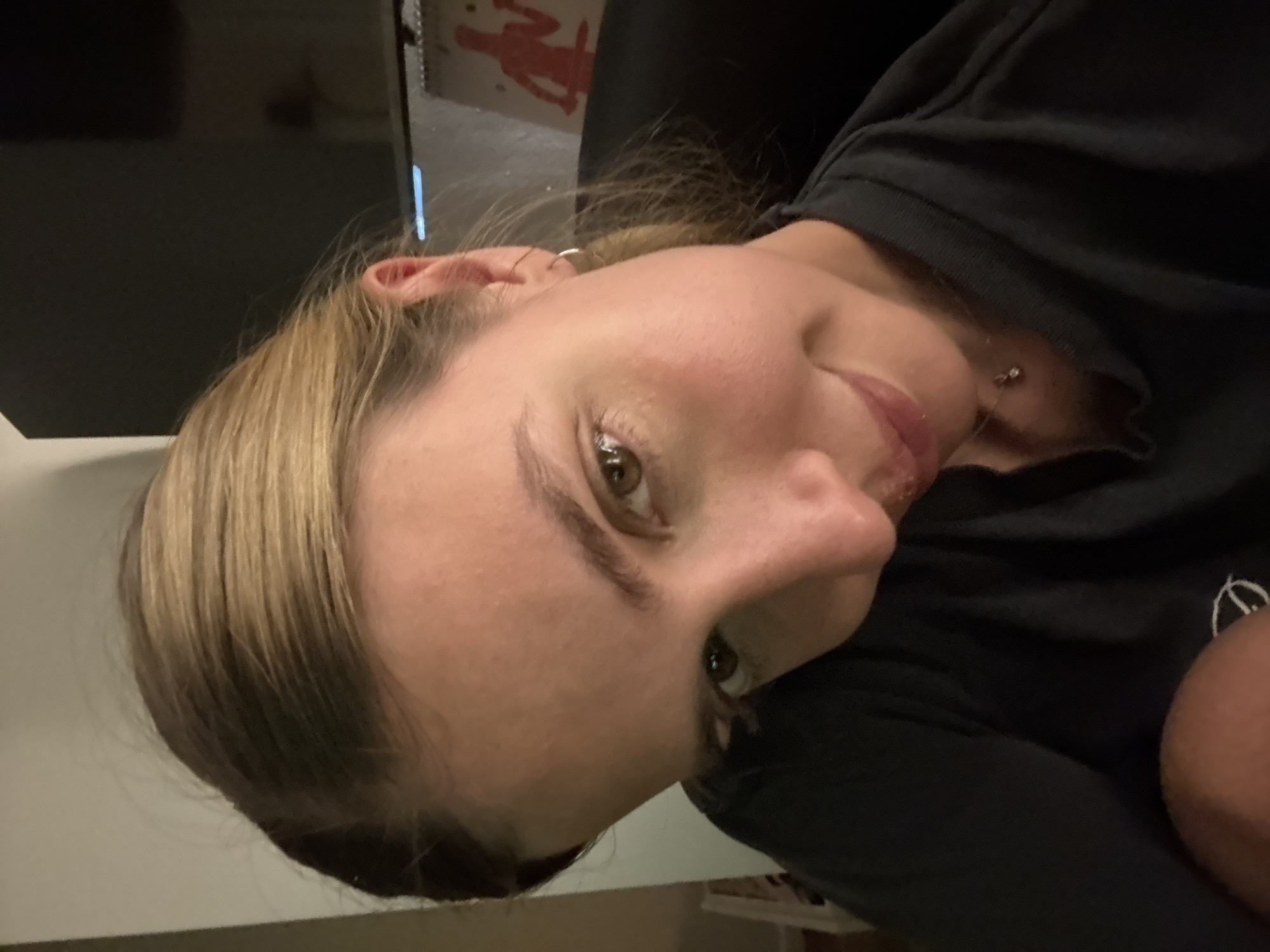 A close-up, medium-shot captures a woman with fair skin and light brown hair. She is wearing a dark-colored shirt. The background is blurred, showing a dark object to the left and a red graphic behind her head. A faint light source illuminates her face from the left. A small piece of jewelry is visible on her neck, and she has a ring in her ear. The scene's location is unknown.