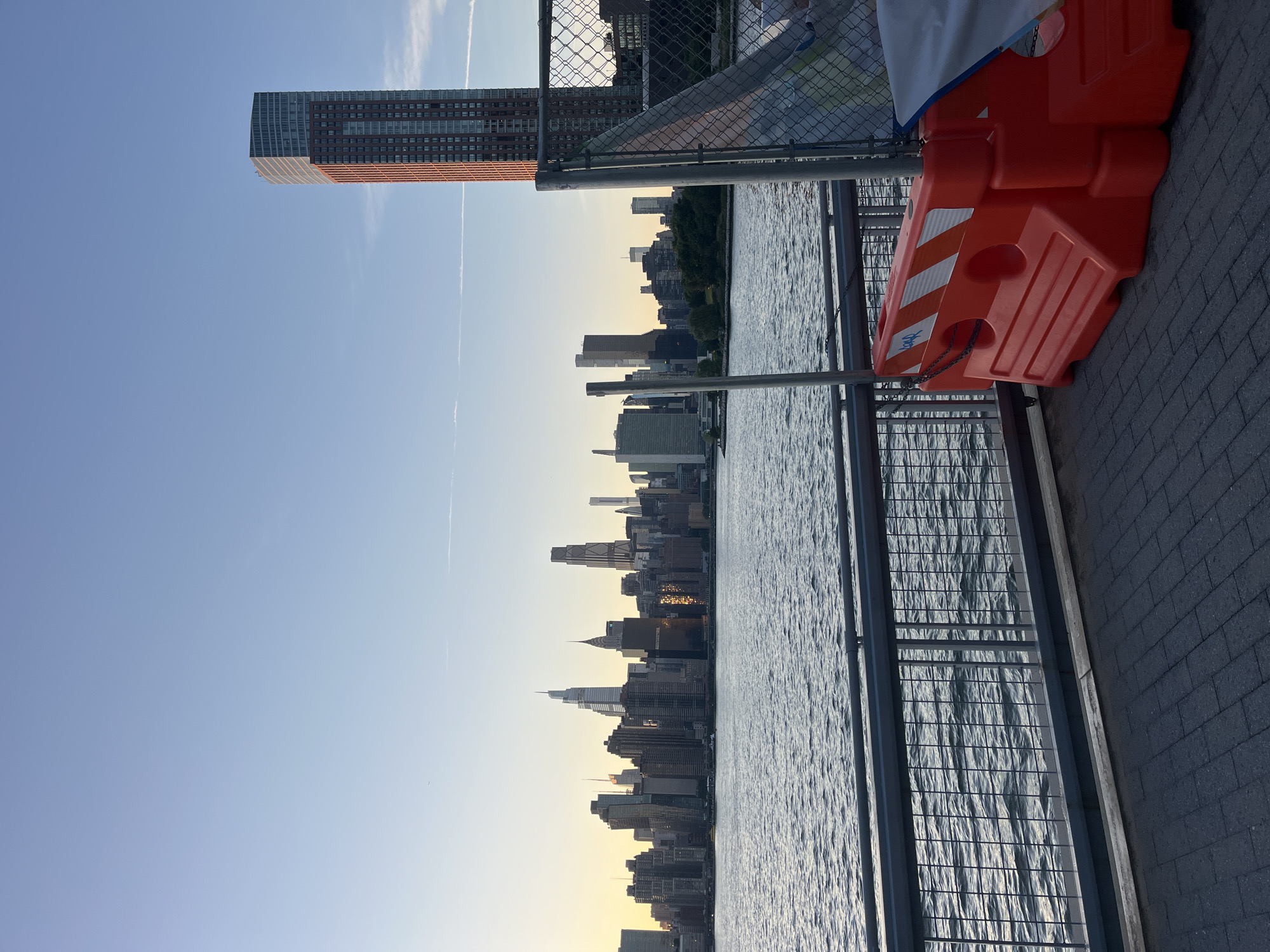 The image depicts a cityscape of New York City, likely from the Queens side looking towards Manhattan, at dusk or early morning. The sky is a gradient of pale blue to soft yellow near the horizon, with a few wispy clouds. A wide body of water, likely the East River, occupies the lower half of the image, with gentle ripples on its surface. A promenade or walkway with a dark, brick-like pavement is in the foreground, separated from the water by a metal railing. To the right of the walkway, a bright orange plastic barrier with reflective white and orange stripes is visible, partially covered by a light-colored tarp. A chain is attached to the barrier and the railing. Beyond the railing and the water, the iconic Manhattan skyline stretches across the horizon, featuring numerous skyscrapers of varying heights and architectural styles. The sun, low in the sky, casts a warm glow on some of the buildings, creating reflections. In the far right of the frame, a tall, reddish-brown skyscraper with many windows stands out against the sky, and a chain-link fence is present in front of it. The overall scene is serene and showcases the grandeur of the city.