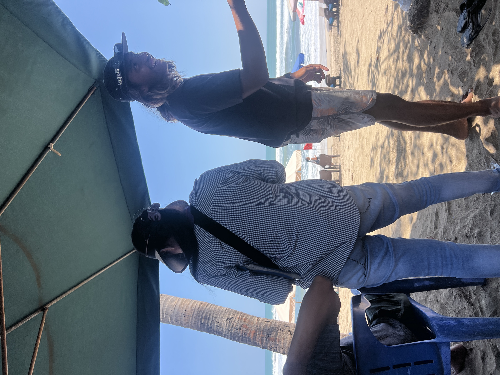 Two individuals are standing on a sandy beach in Kuta, Indonesia, under a large green umbrella. The person on the left, viewed from the back, wears a black hat and a blue and white checkered long-sleeved shirt with a black strap over their shoulder. They are also wearing light blue jeans. To the right, a person wearing a black baseball cap with "ANGELS" written on it and sunglasses on the brim, is smiling and looking to the right. They are wearing a black t-shirt and patterned shorts. The background shows the ocean with waves, beach chairs, and umbrellas. A palm tree is visible to the left. The scene appears to be a casual beach setting.