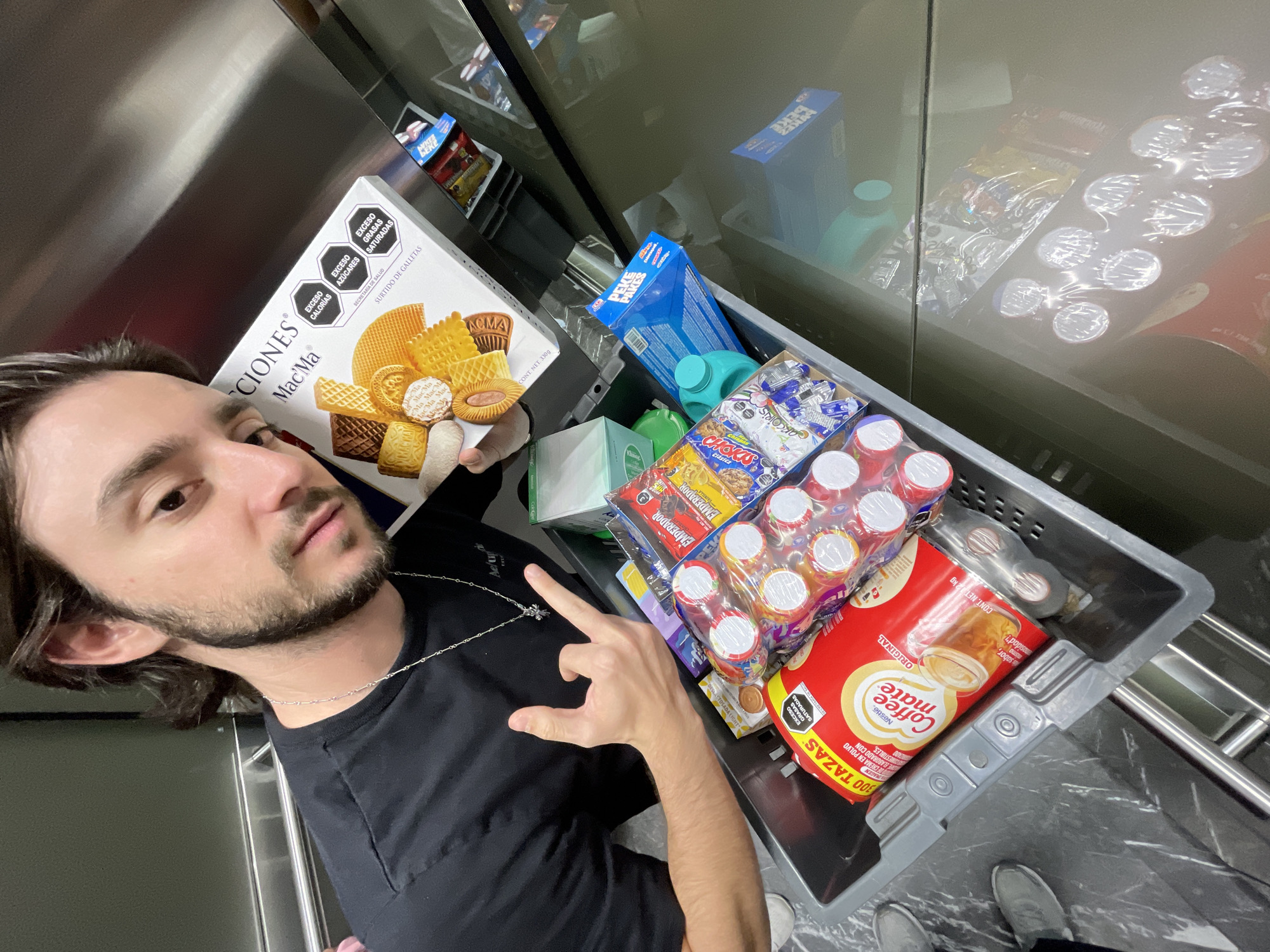 A waist-up shot shows a man inside a supermarket. He has brown hair, a light beard, and a silver chain, wearing a black T-shirt. The man is holding a box of assorted cookies from Mac’Ma. His left hand is raised, forming the rock-on symbol.
Next to him is a gray shopping basket full of groceries. The items include a box of Pekke Pakkes cookies, a pack of Emperador cookies, Nesle Coffee Mate, and various beverages in small plastic bottles.
The supermarket is a well-lit area with shelves stocked with food. The setting indicates a typical shopping trip in Mexico City, Mexico, given the products are labeled in Spanish and contain OCTA tags.