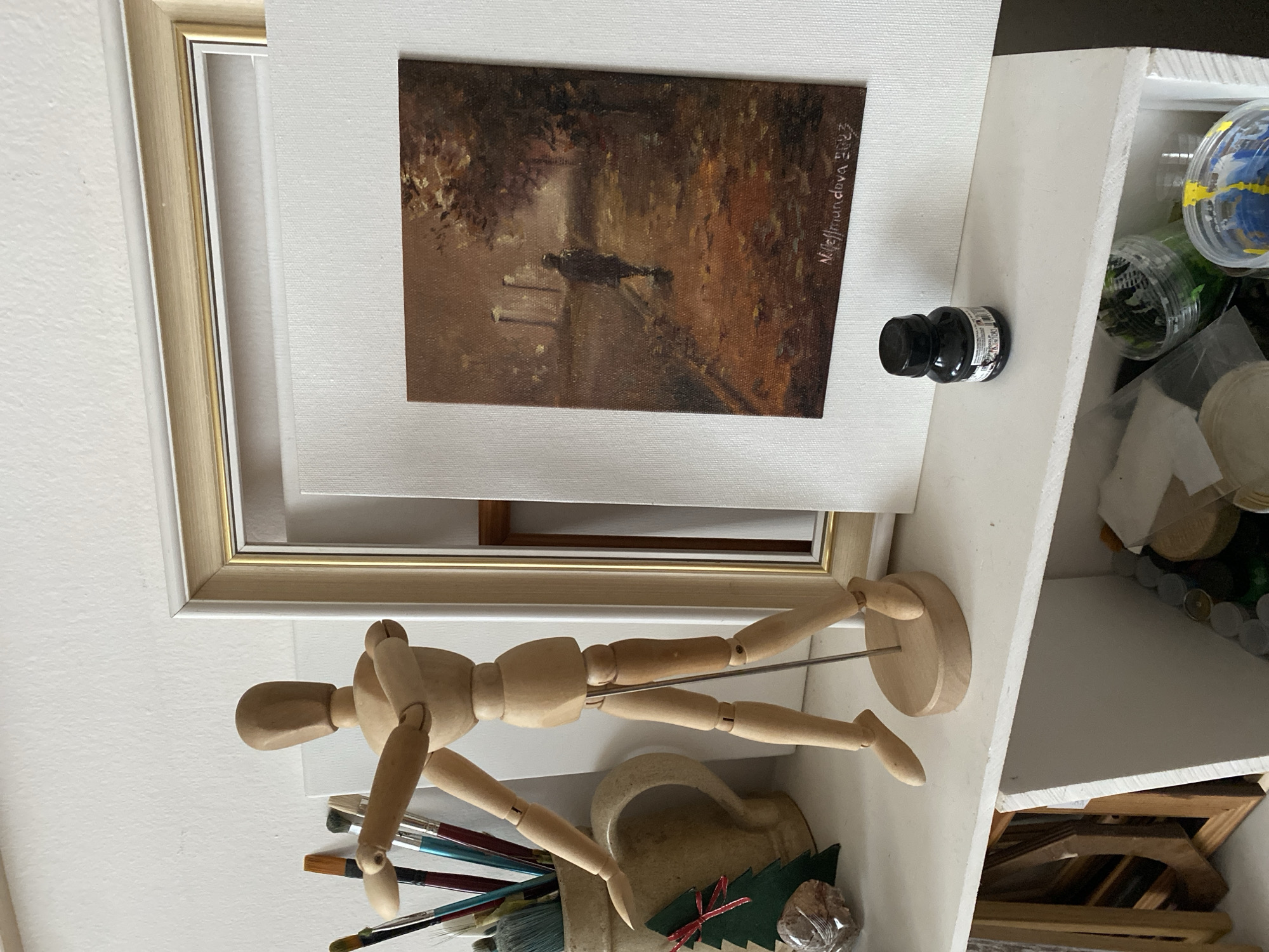 A wooden artist's mannequin stands on a white shelf to the left of a framed painting of a solitary figure walking down a dimly lit street at dusk. To the left of the mannequin are several paintbrushes in a ceramic jug, a small decorative Christmas tree, and a rock. To the right of the painting is a small black bottle of ink. The shelf is filled with art supplies and other miscellaneous items. The background is a white textured wall. There is no indication of the location being the city of Bílina, Czechia.