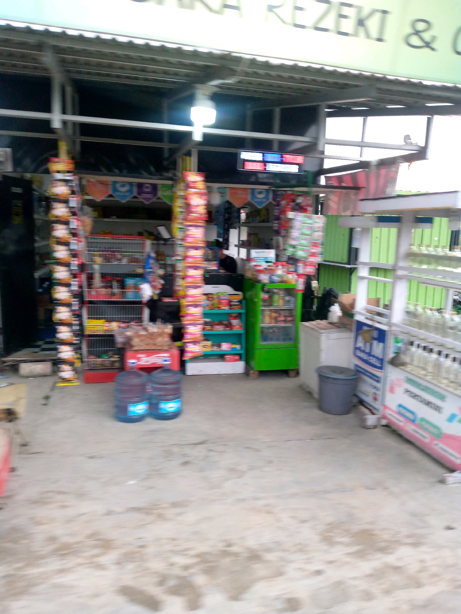 The image depicts the exterior of a small convenience store, likely a warung, in Keudenibong, Indonesia. The storefront features a covered awning with a green and white sign that is partially legible, showing "REZEKI &". Shelving units are visible, stocked with various goods, including bottled drinks, snacks, and what appear to be pet food or supplies in cages. A green refrigerator unit is also present, displaying drinks and food items. In front of the store, two large blue water jugs are placed on the ground. The environment appears to be a street with a concrete surface. No people are clearly visible in the scene. The image is taken from a medium distance, with some motion blur present.