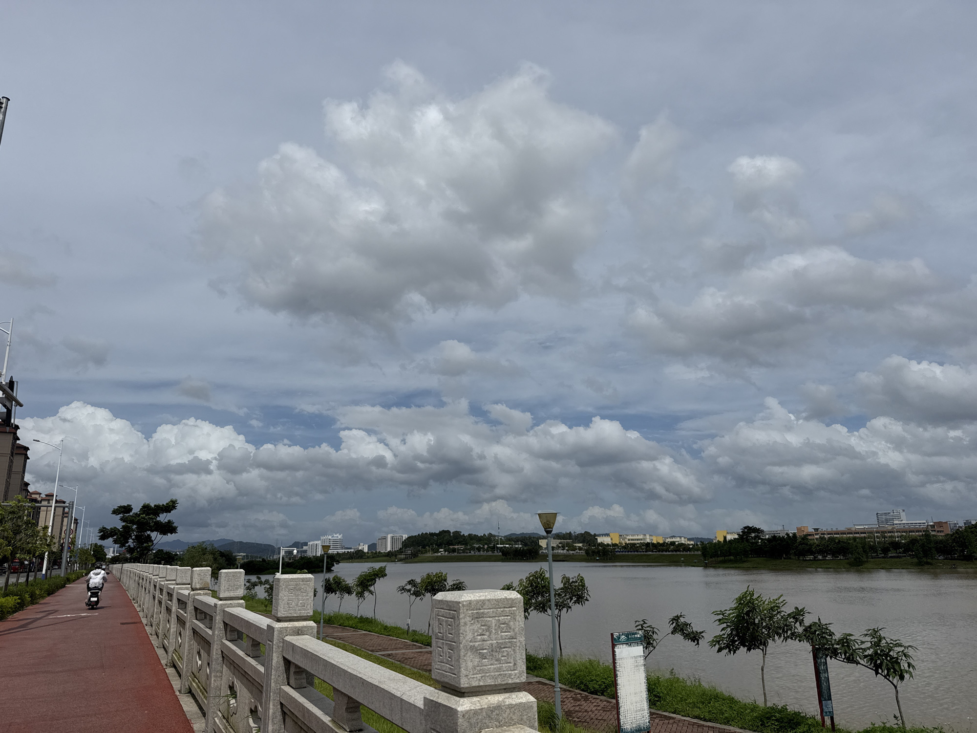 This media file depicts a scenic view of a waterfront promenade in Huizhou, China. The scene is dominated by a wide, calm body of water on the right, reflecting the overcast sky. On the left, a red paved pathway runs alongside a stone railing, suggesting a recreational area. A person on a scooter is moving away from the viewer on this pathway.
The sky is filled with large, dramatic cumulus clouds, indicating a cloudy or potentially stormy day, likely mid-morning or early afternoon given the ambient light. The setting appears to be an urban or suburban area, with buildings visible in the distance across the water, along with trees and a gentle slope of land. The stone railing features decorative carvings, adding an artistic element to the infrastructure.
The overall mood is serene, with the vastness of the sky and water providing a sense of peace, contrasted with the movement of the scooter. There is no discernible text in the visible portion of the image. The location context of Huizhou, China, suggests a potentially tropical or subtropical climate, which could contribute to the dramatic cloud formations.