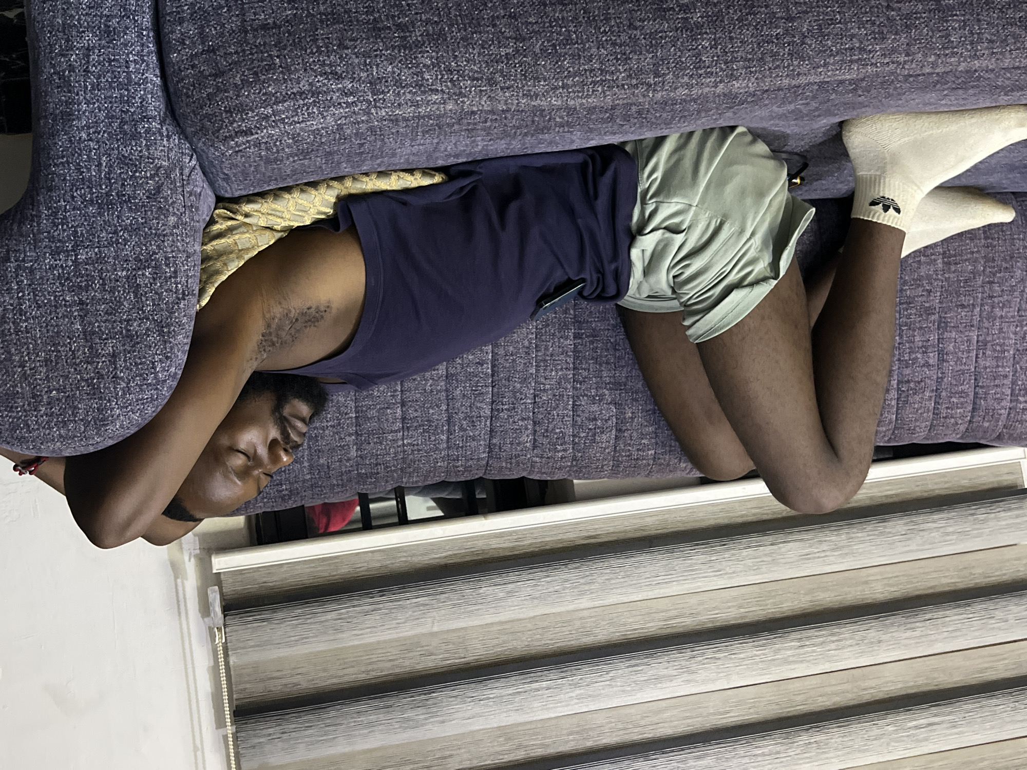 A dark-skinned man is asleep on a purple couch in what appears to be a residential setting, possibly in Abuja, Nigeria. He is wearing a navy blue tank top, light green shorts, and white socks with the Adidas logo. His head is resting near the arm of the couch and his body is oriented parallel to a closed window blind. A gold-colored cushion is visible behind the man.