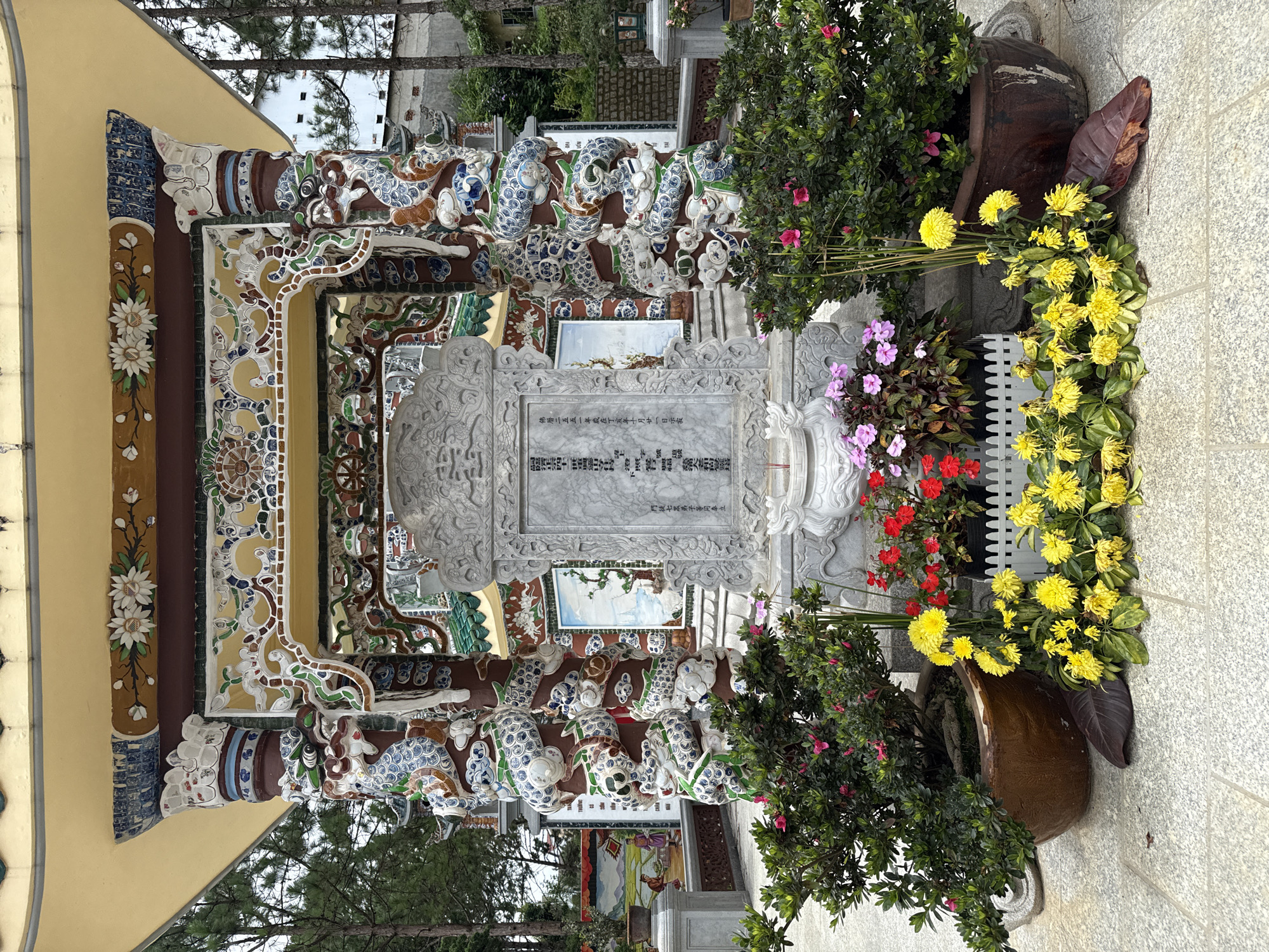 The image captures a detailed view of a monument located within the grounds of the Minh Mang Tomb in Da Lat, Vietnam. The monument, a large, ornate stone stele, is intricately carved and surrounded by decorative architectural elements. Flanking the stele are two large, brown ceramic pots overflowing with lush green foliage and vibrant pink flowers. In front of the monument, a decorative white picket fence encloses a cluster of bright yellow chrysanthemums, some of which appear slightly wilted, and smaller red and pink flowers. The background reveals further elements of the tomb's architecture, including ornate pillars adorned with colorful dragon motifs, tiled roofs, and painted murals depicting traditional scenes. The overall scene is one of solemn beauty, blending natural elements with elaborate historical artistry, set against a backdrop of evergreen trees characteristic of the Da Lat region.