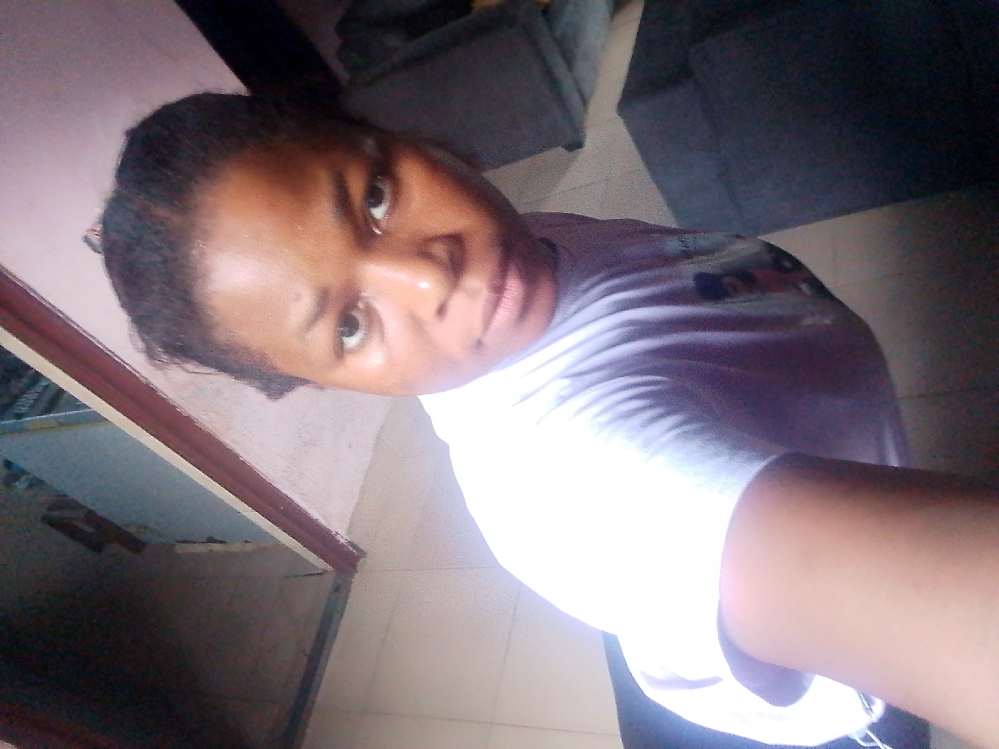 The image shows a young Black woman taking a selfie. She is wearing a light purple t-shirt with a subtle pattern. Her hair is dark and pulled back. Her facial expression appears neutral or slightly inquisitive, with her eyes looking directly at the camera.
The setting appears to be indoors, with a tiled floor visible. To the left of the frame, there's a wooden structure, possibly a doorway or cabinet, and beyond that, a glimpse of what could be furniture or a shelf. In the background to the right, a dark, plush surface is visible, suggesting a sofa or armchair. The lighting is somewhat dim, with a strong light source casting a bright glow on the woman's shoulder and part of her shirt, suggesting the flash from her device or a nearby light.
There is no visible text in the image. The overall mood is informal and personal, typical of a selfie. Without further context, the exact activity or event is unclear, but it appears to be a casual moment captured by the subject. The location is specified as Yenagoa, Nigeria, which is a context clue but not directly observable from the image itself.